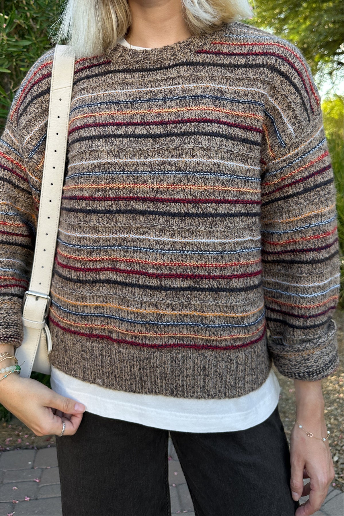 A woman wearing a brown and beige striped knit sweater with horizontal multicolor stripes, styled with a white undershirt, black pants, and a cream shoulder bag, set against an outdoor background of green foliage.
