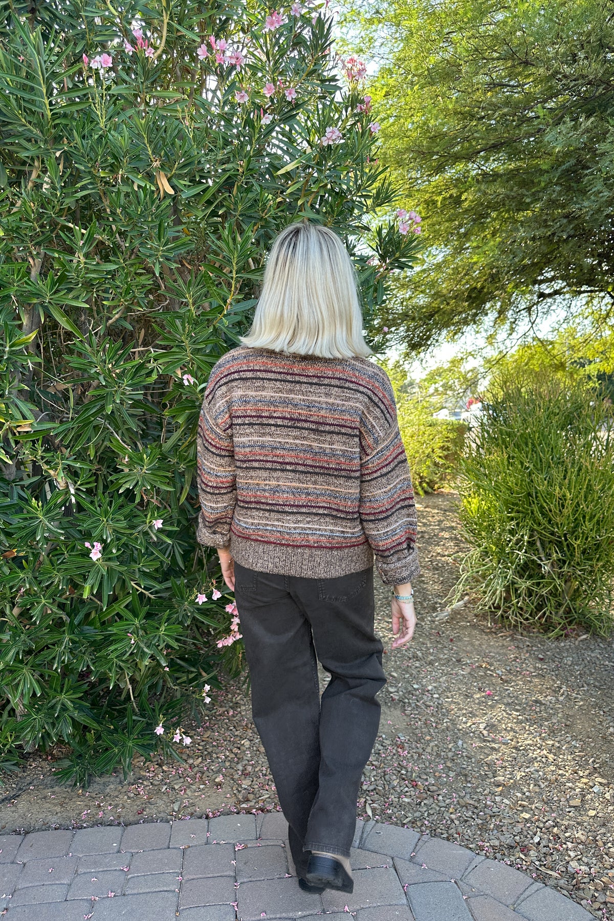 A woman with blonde hair is seen from behind, wearing a striped knit sweater and dark trousers as she walks along a paved garden path surrounded by green bushes and trees.