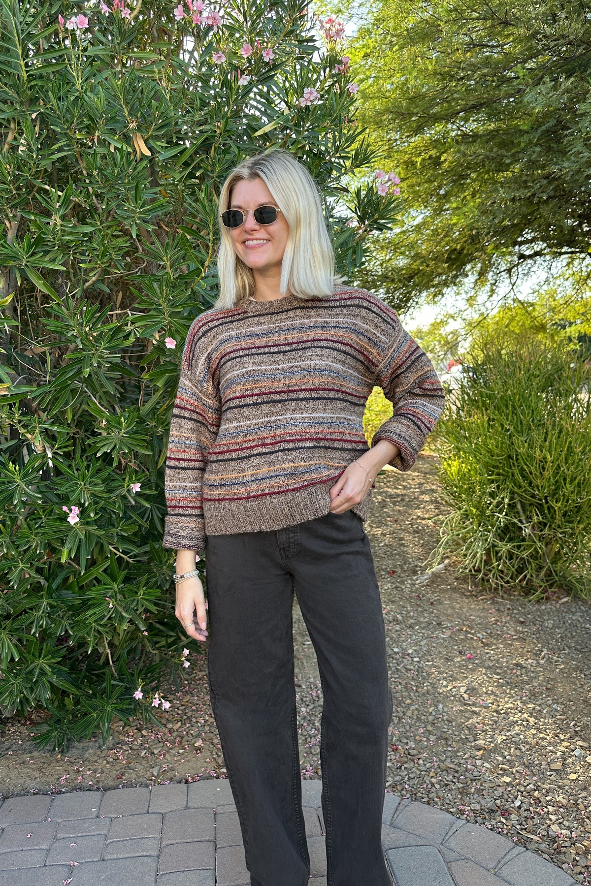 A woman with blonde hair and sunglasses stands outdoors in a garden setting, wearing a long-sleeve knit sweater with horizontal stripes in brown, beige, and muted red, paired with dark charcoal pants.