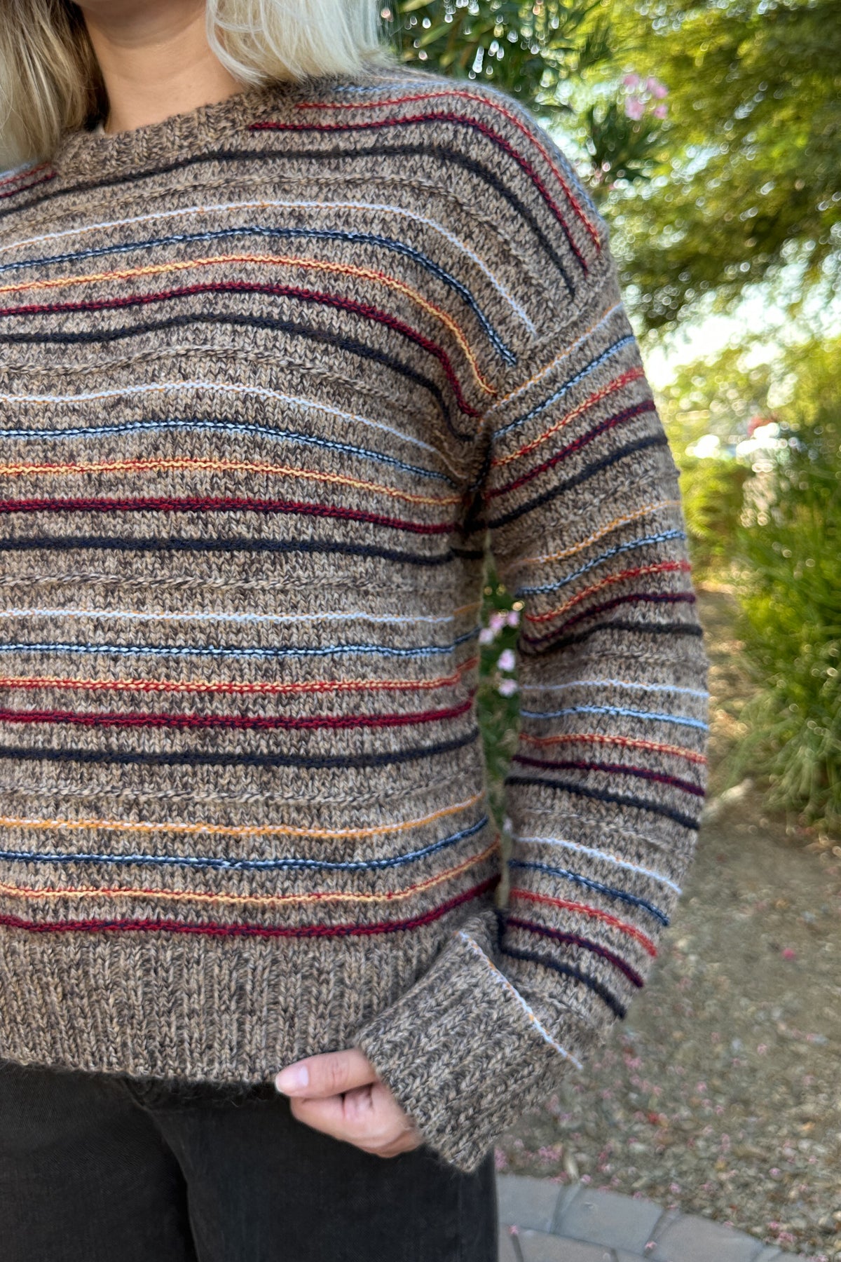 A close-up of a person wearing a chunky knit sweater with horizontal stripes in brown, beige, orange, blue, and burgundy, photographed outdoors with a blurred garden background.