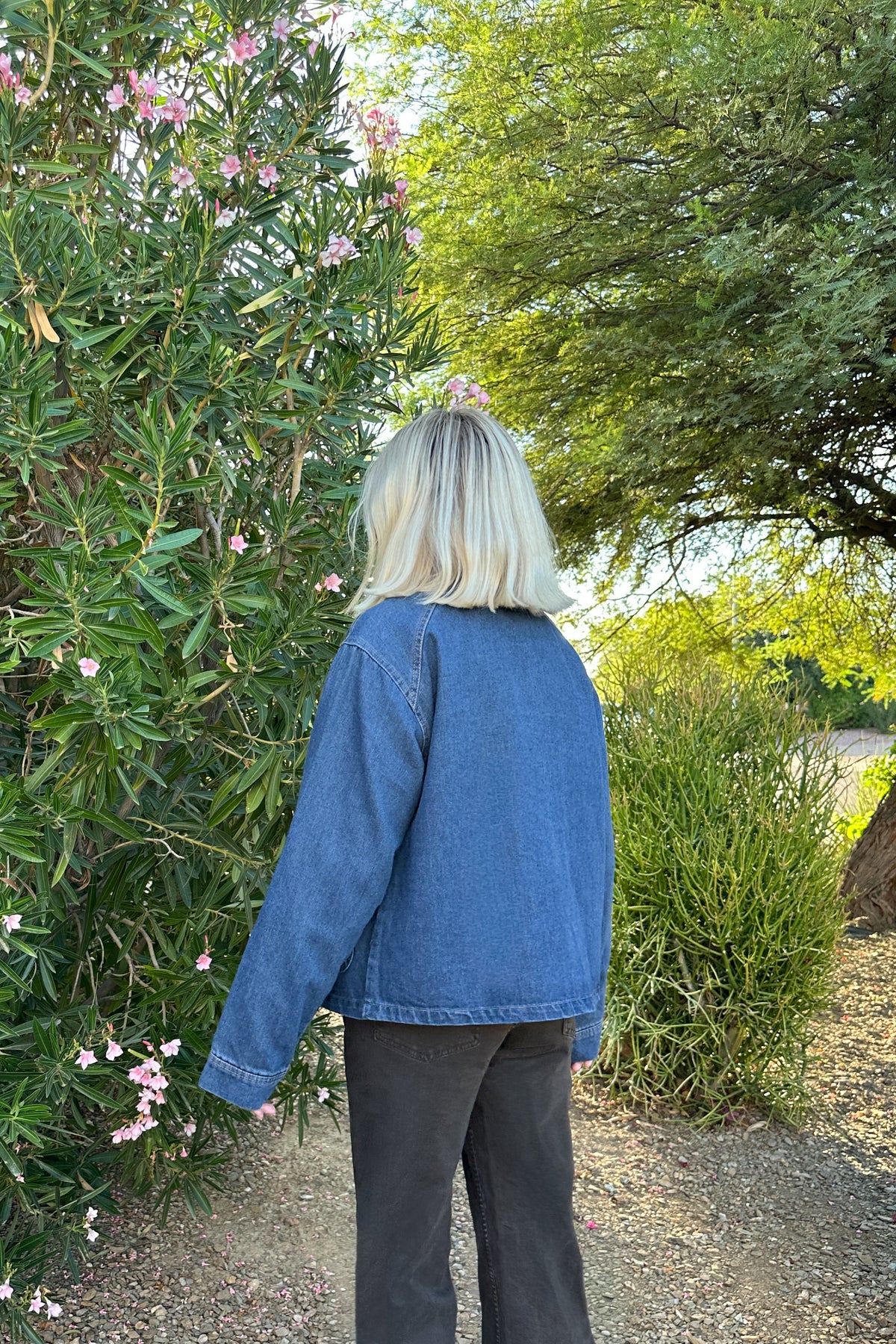 A person viewed from behind wearing a blue denim jacket and dark gray pants, standing outdoors in a garden next to green bushes with pink flowers.