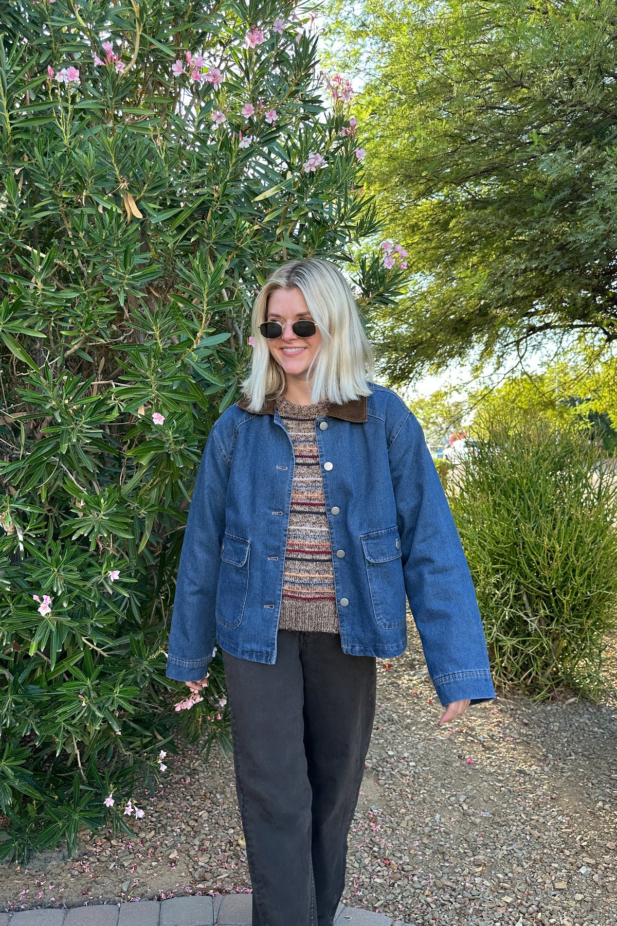 A woman wearing a blue denim jacket with a brown corduroy collar, a patterned knit top, and black pants, standing outdoors in front of green bushes and trees.