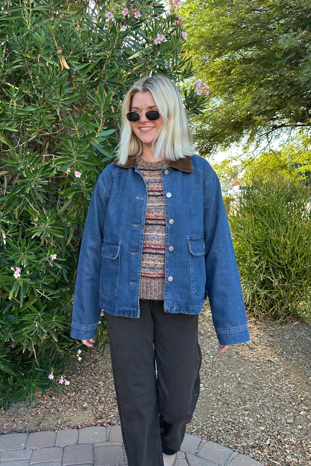 A woman wearing a blue denim jacket with a brown corduroy collar, a patterned knit top, and black pants, standing outdoors in front of green bushes and trees.