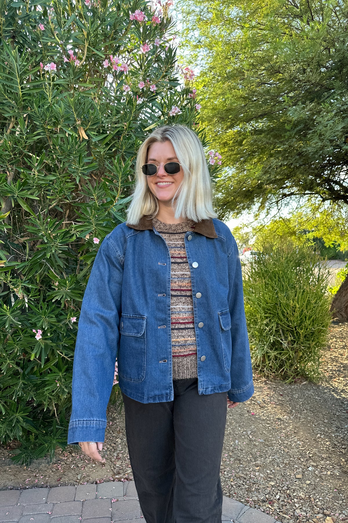 A woman wearing a blue denim jacket with a brown corduroy collar, a patterned knit top, and black pants, standing outdoors in front of green bushes and trees.