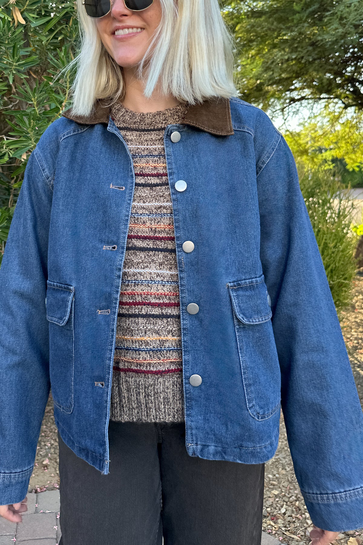 A woman wearing a blue denim jacket with a brown corduroy collar, a patterned knit top, and black pants, standing outdoors in front of green bushes and trees.
