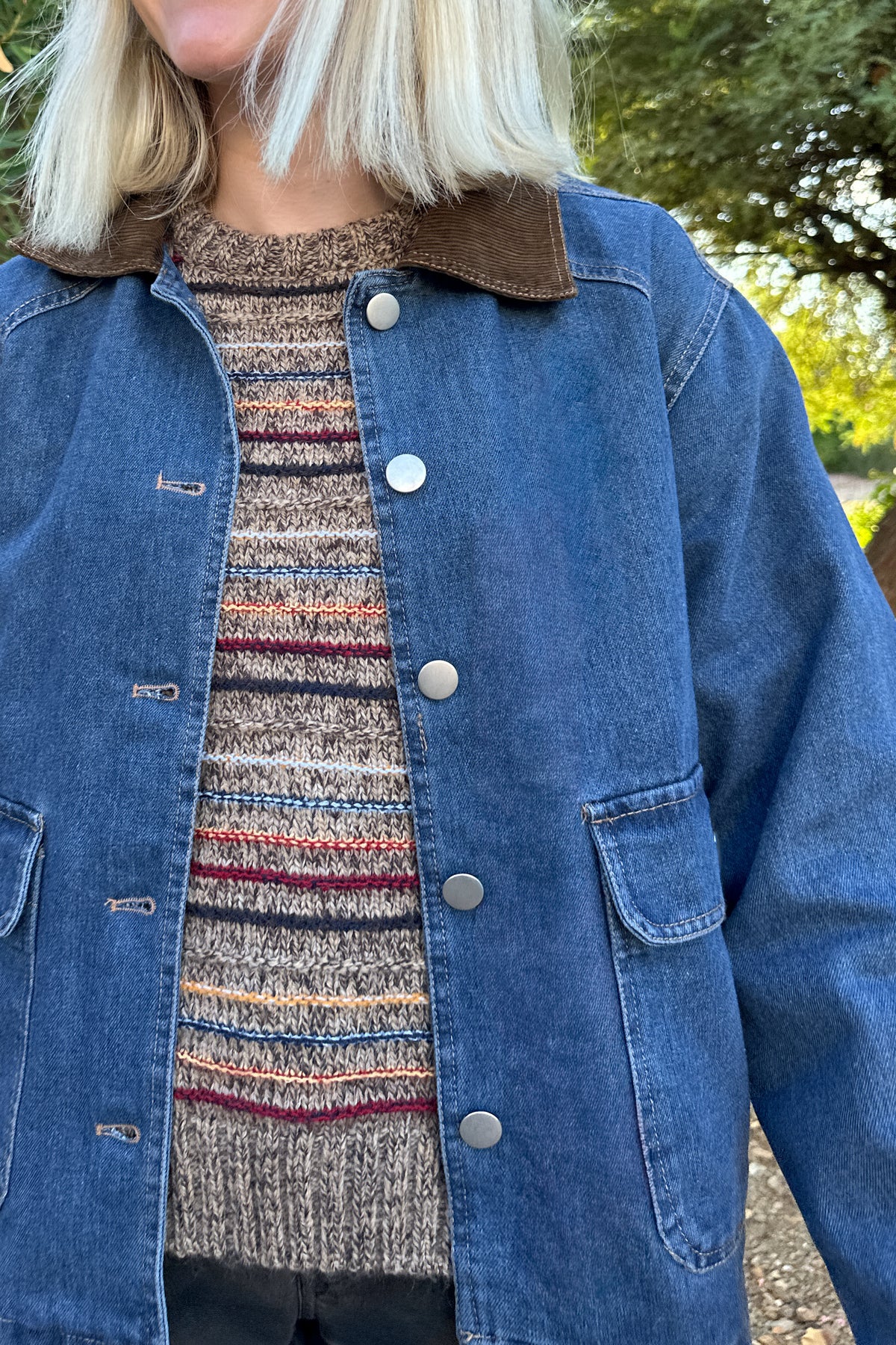 A person wearing a blue denim jacket with a brown corduroy-style collar over a striped knit sweater, photographed outdoors with blurred greenery in the background.