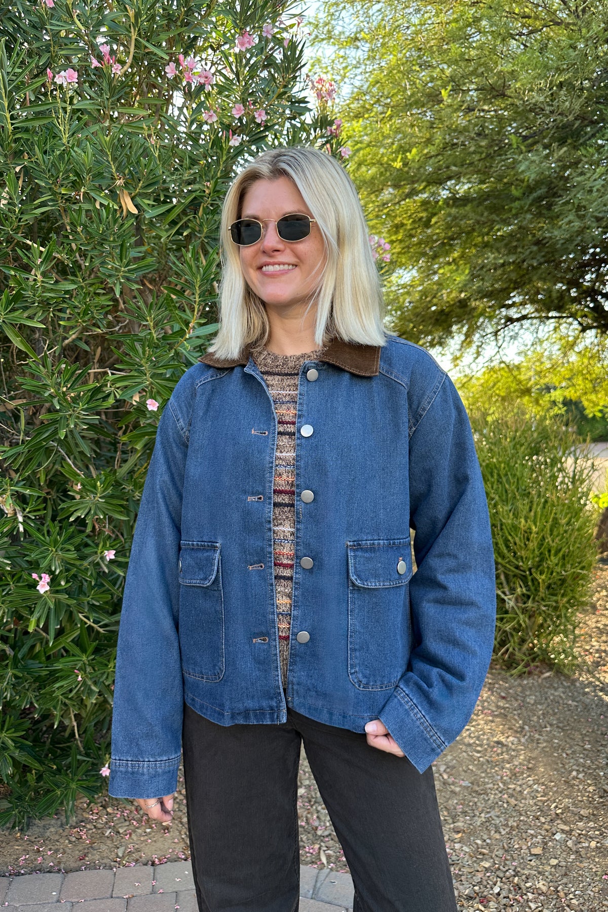 A woman wearing a blue denim jacket with a brown corduroy collar, a patterned knit top, and black pants, standing outdoors in front of green bushes and trees.
