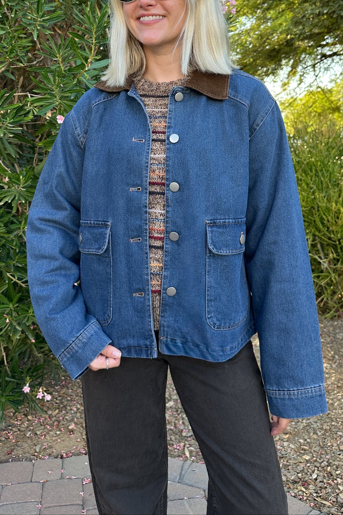 A woman wearing a blue denim jacket with a brown corduroy collar, a patterned knit top, and black pants, standing outdoors in front of green bushes and trees.