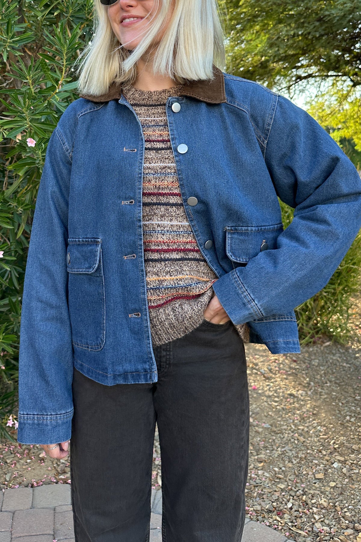 A woman wearing a blue denim jacket with a brown corduroy collar, a patterned knit top, and black pants, standing outdoors in front of green bushes and trees.