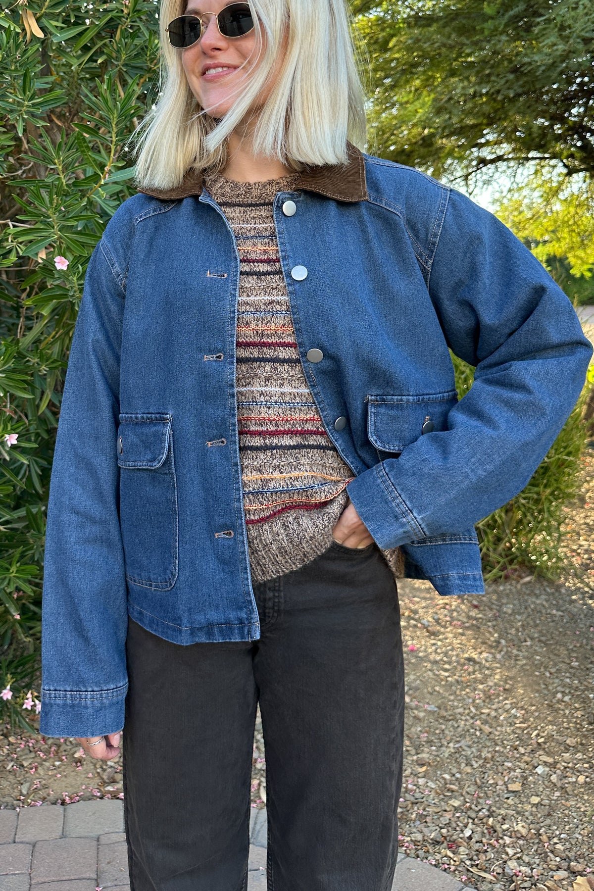 A woman wearing a blue denim jacket with a brown corduroy collar, layered over a striped knit sweater and black pants, standing outdoors in front of green foliage.