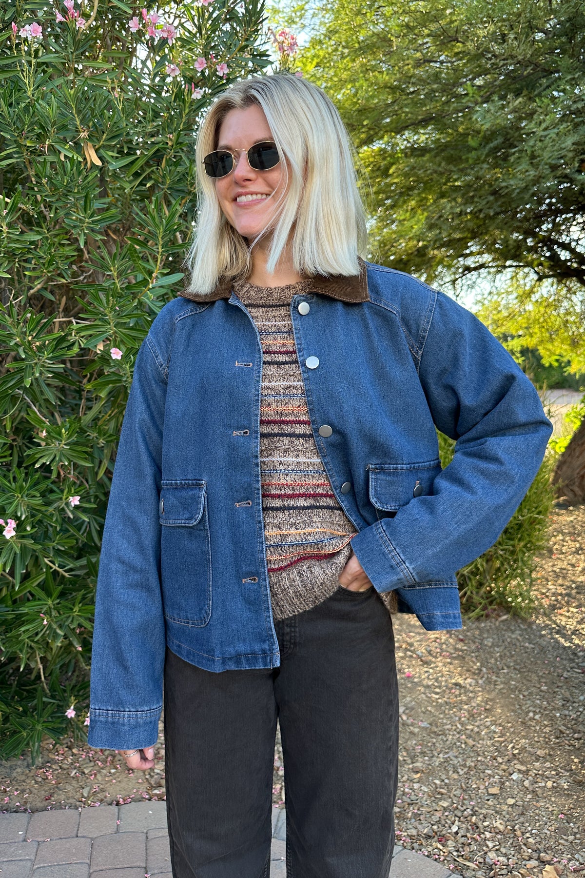 A woman wearing a blue denim jacket with a brown corduroy collar, a patterned knit top, and black pants, standing outdoors in front of green bushes and trees.
