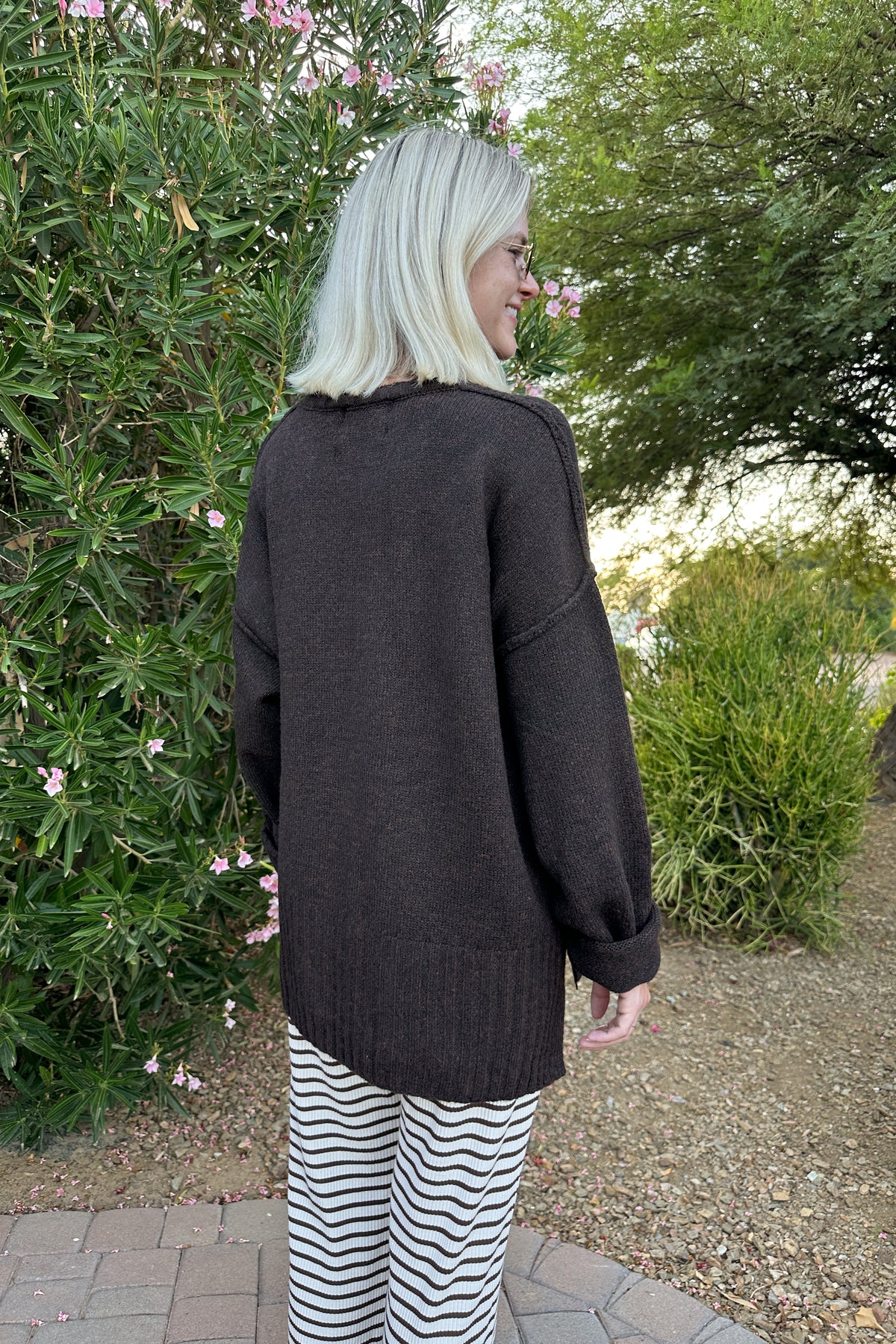 A woman with platinum blonde hair is shown from behind wearing a dark brown oversized knit sweater and black and white striped pants, standing outdoors in a garden setting.
