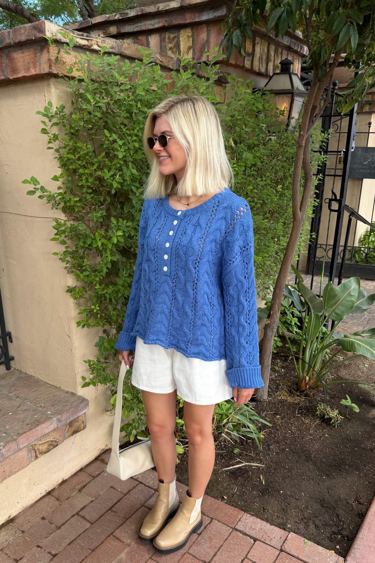 A woman stands outdoors wearing a blue cable-knit long-sleeve top with white buttons, white shorts, beige ankle boots, and carrying a cream shoulder bag.