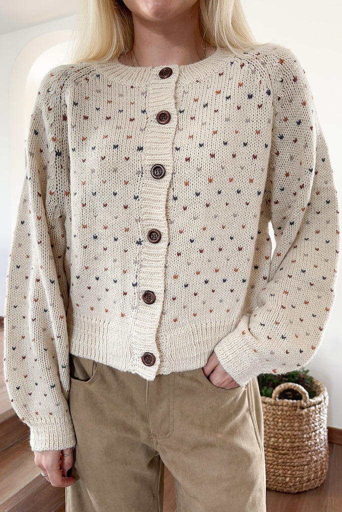 Person wearing a beige knitted cardigan with small patterns and brown buttons, standing indoors.