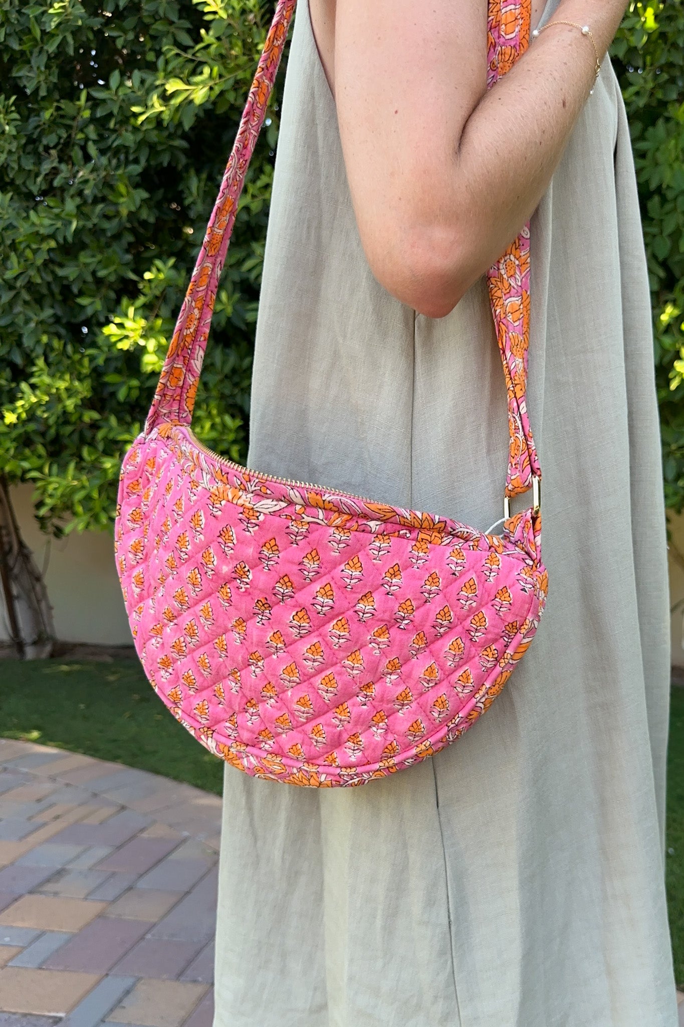 A pink quilted crescent shoulder bag with a small floral pattern and a matching patterned strap, worn by a person in a light beige dress outdoors.