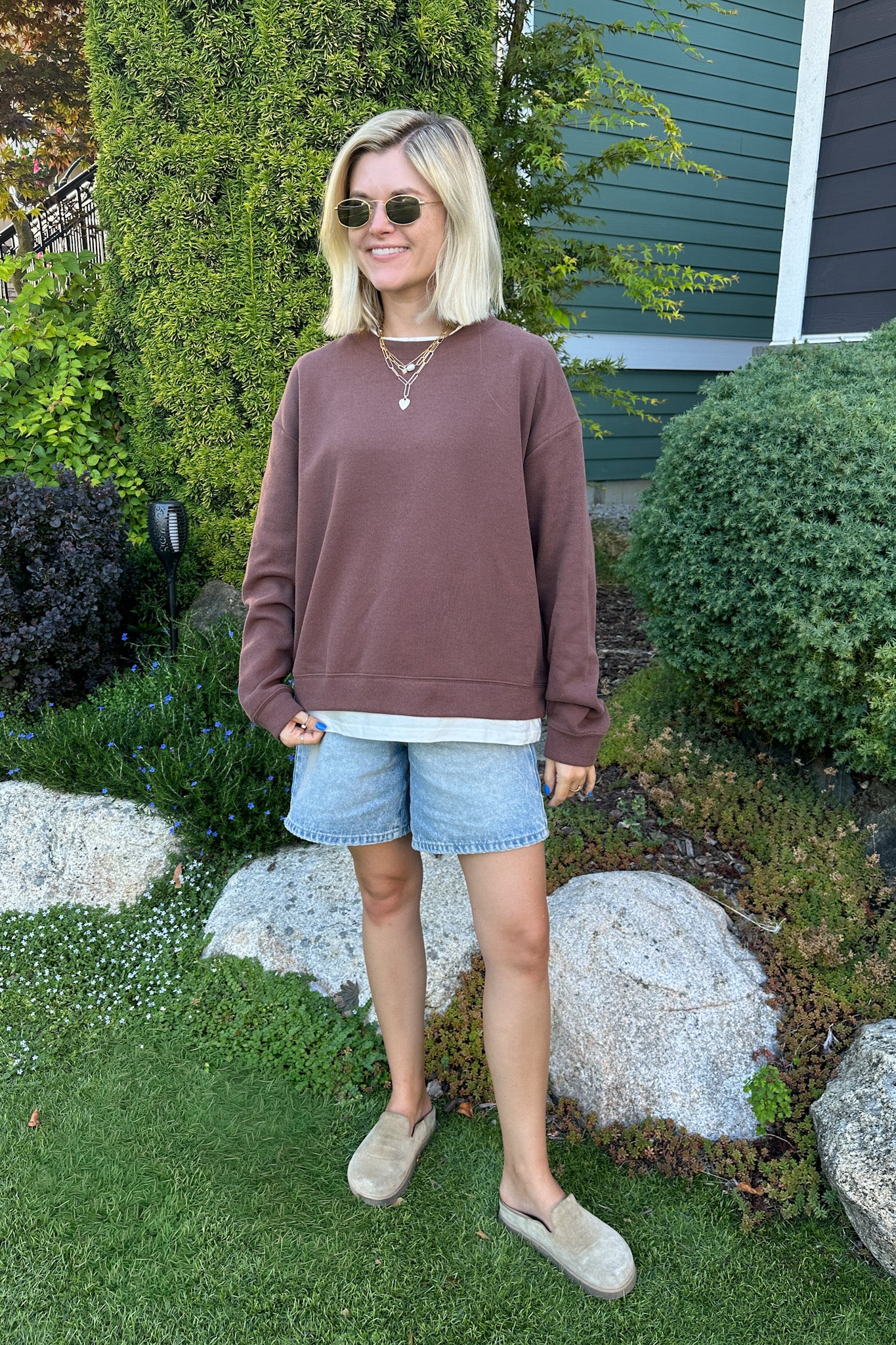 A woman wearing a dusty rose-brown crewneck sweatshirt, denim shorts, and beige slip-on shoes stands in a landscaped garden setting with green grass, shrubs, and large rocks.