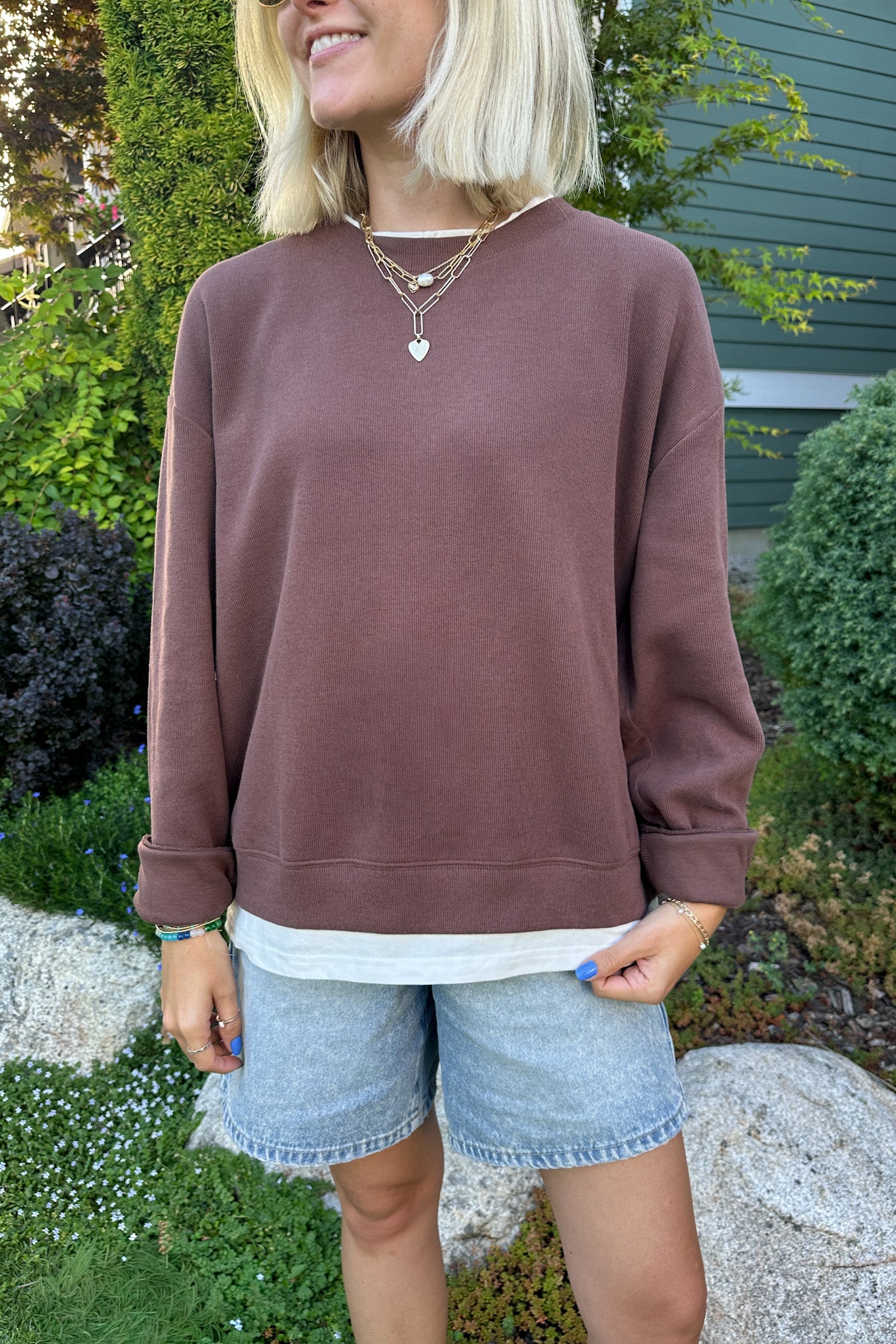 A woman wearing a dusty rose brown crewneck sweatshirt and light-wash denim shorts, standing outdoors in front of green garden plants and a house wall.