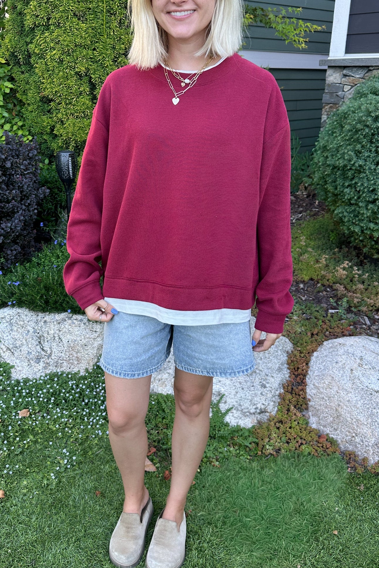 A woman wearing an oversized burgundy crewneck sweatshirt, light blue denim shorts, and beige slip-on shoes, standing in a grassy backyard garden.