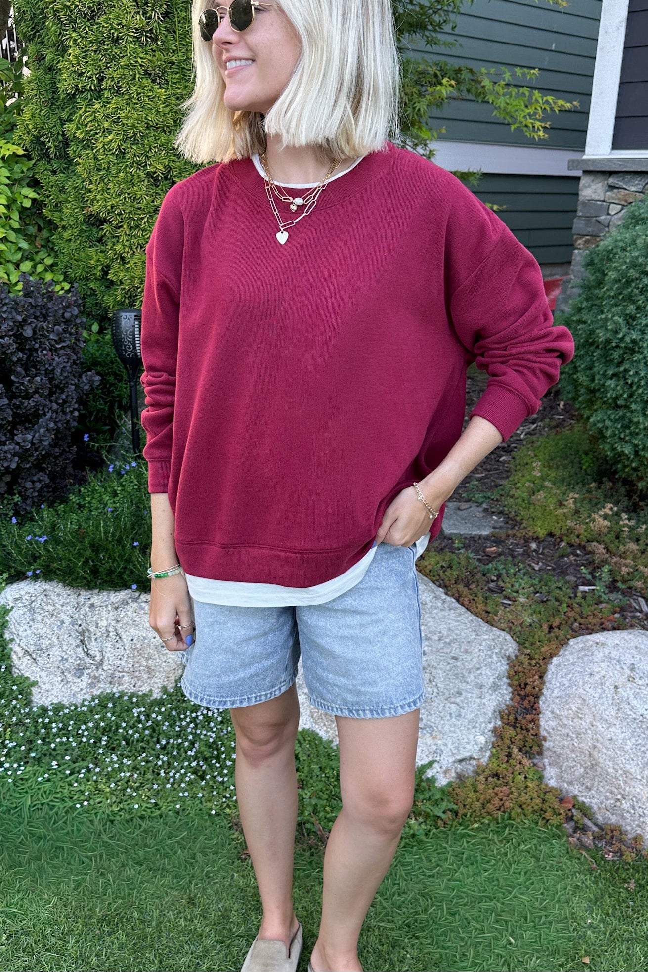 A woman wearing a burgundy crewneck sweatshirt, light-wash denim shorts, and sunglasses, standing outdoors in a grassy yard with garden plants and a house in the background.