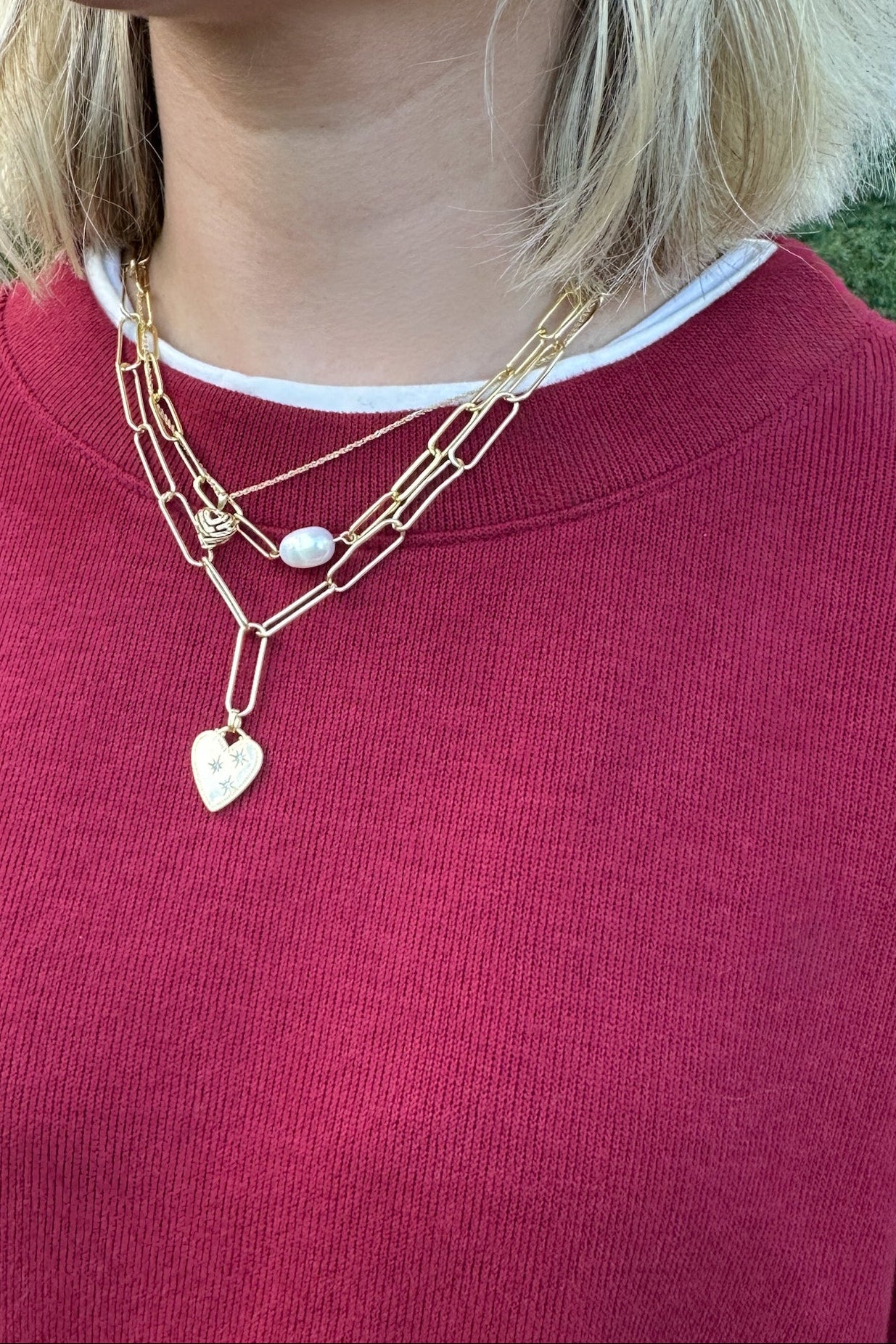 A close-up of a person wearing a layered gold necklace with a heart-shaped pendant and a small white bead, paired with a burgundy sweatshirt.