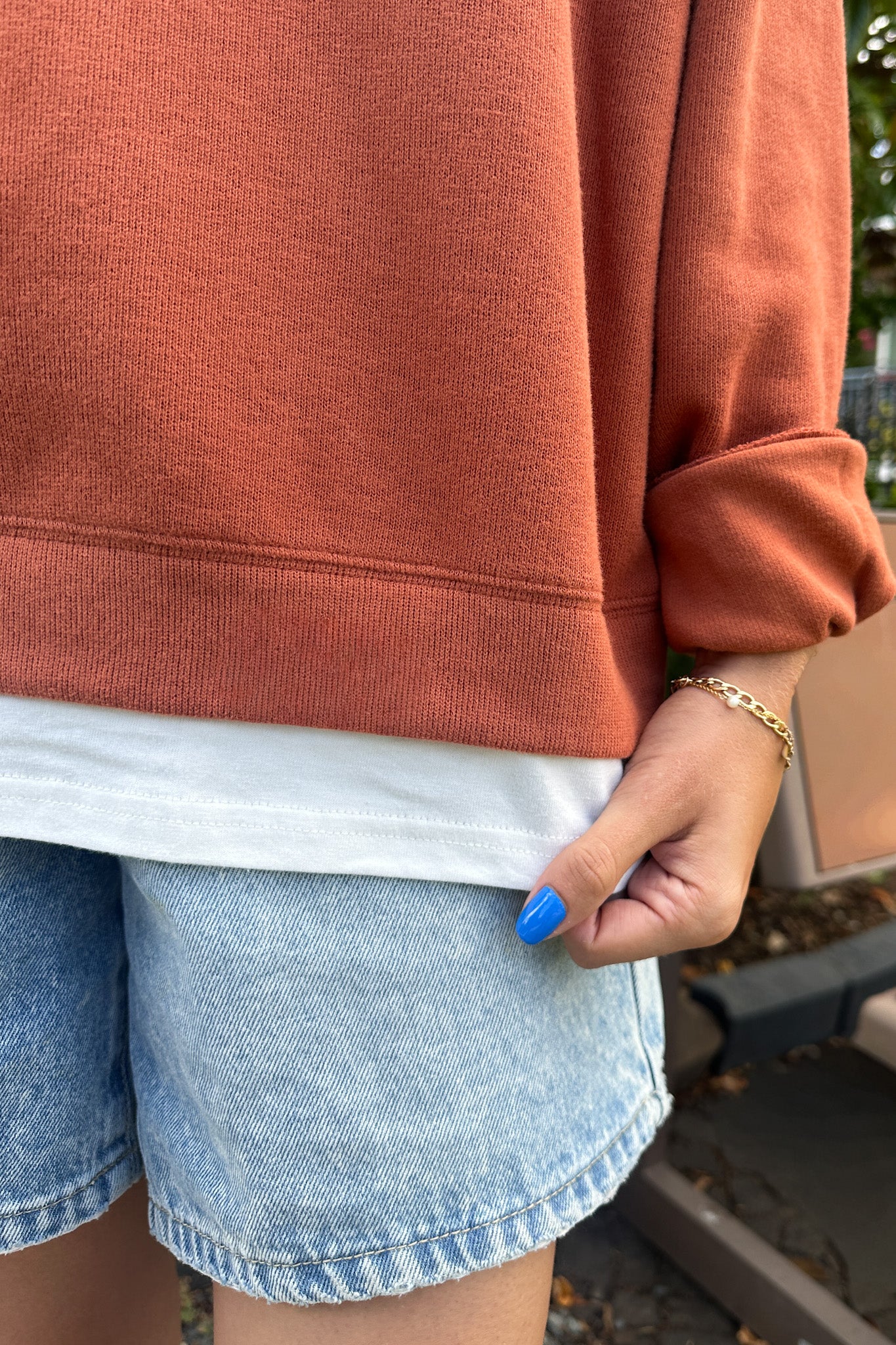 Close-up of a person wearing a rust-orange sweatshirt, a white t-shirt peeking out, and light-wash denim shorts, with a gold chain bracelet on the wrist.