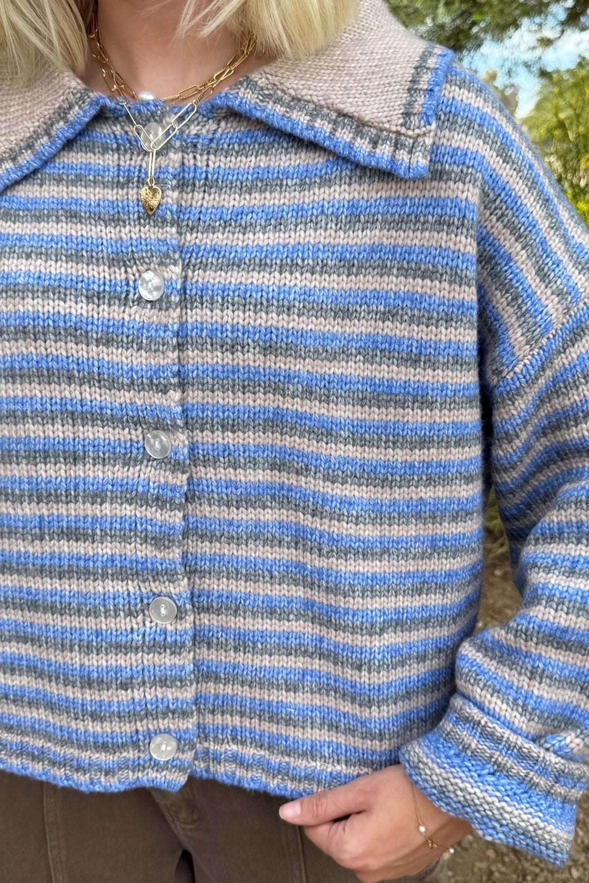A close-up of a person wearing a hand-knit cardigan with horizontal stripes in light blue, gray, and white, featuring a beige knit collar and silver buttons.