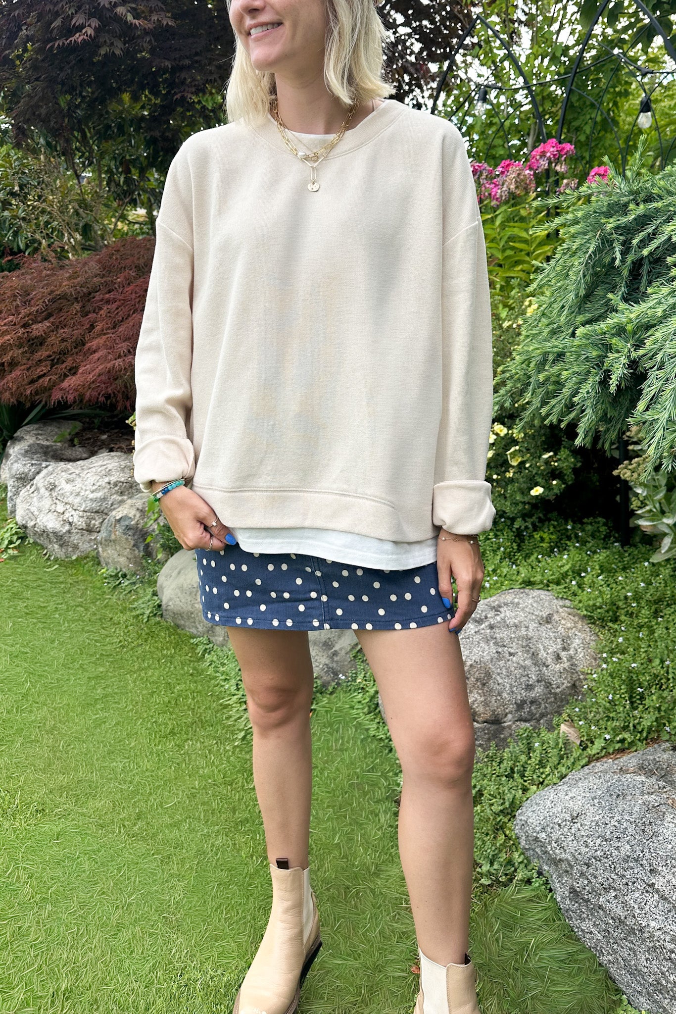 A woman wearing a cream-colored crewneck sweatshirt and a navy blue mini skirt with white polka dots, standing in a garden setting with grass, rocks, and greenery.