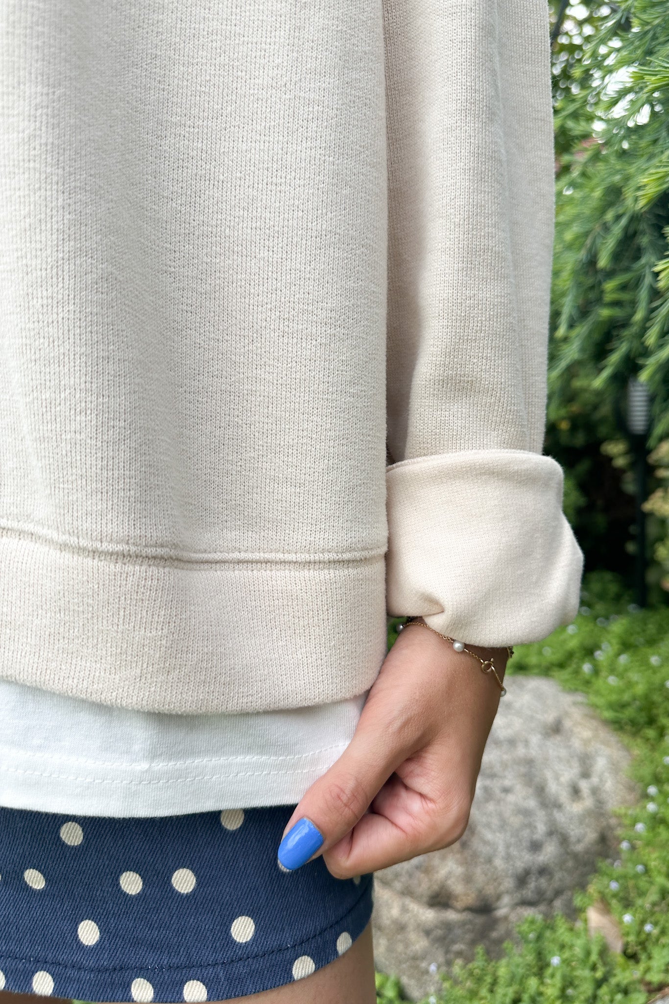 A close-up of a person wearing a cream-colored crewneck sweatshirt with a white undershirt and navy blue polka dot shorts, standing outdoors in front of green foliage.