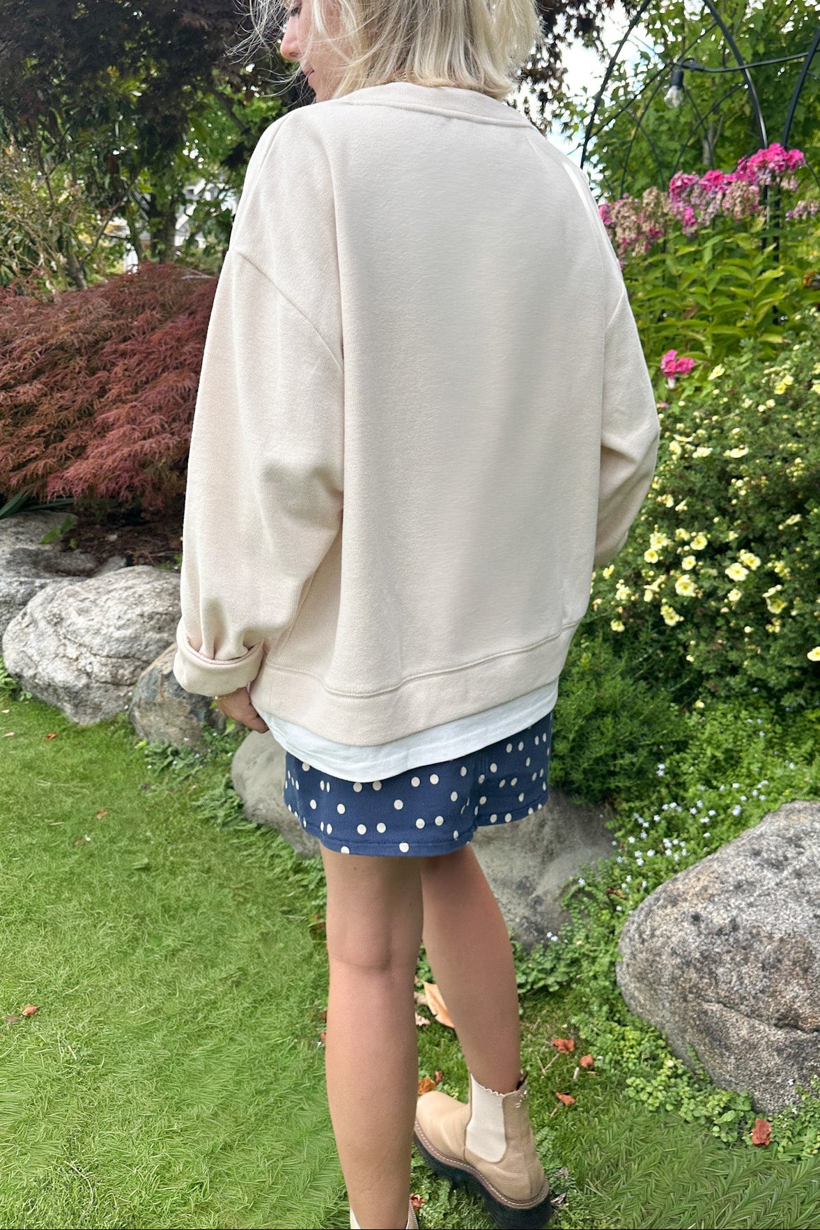 A woman standing outdoors in a garden, viewed from behind, wearing an oversized cream sweatshirt, a navy blue polka-dot skirt, and beige ankle boots.
