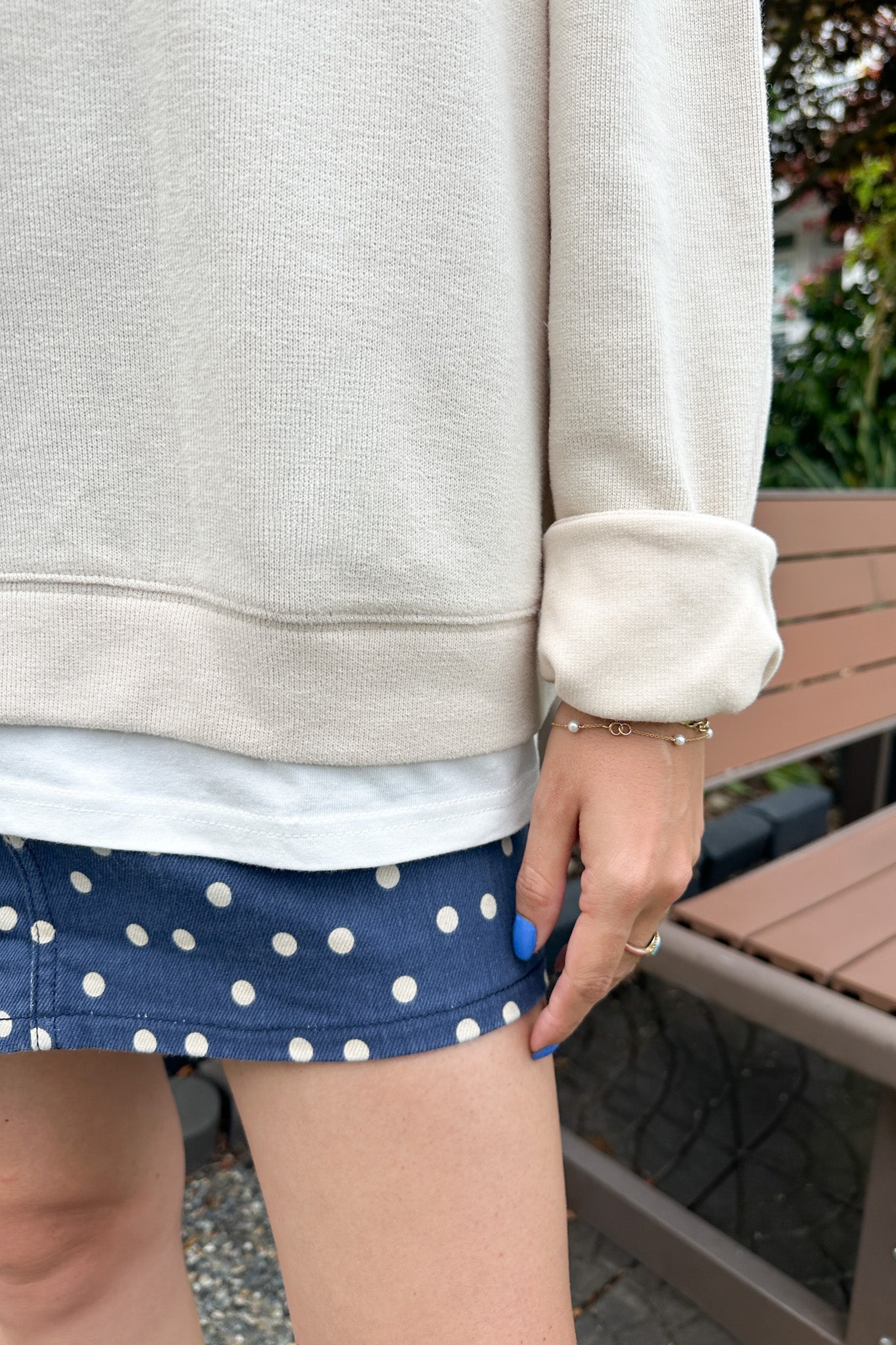 A person outdoors wearing a cream-colored sweatshirt, a white t-shirt, and navy blue shorts with white polka dots. A wooden park bench and green foliage are visible in the background.