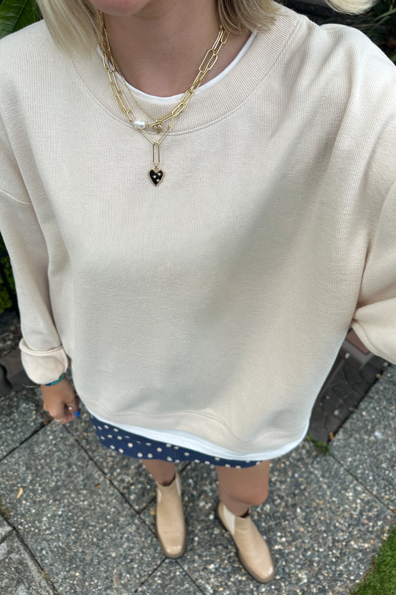 A person wearing an oversized cream-colored ribbed sweatshirt, layered with gold chain necklaces, a navy blue polka-dot skirt, and beige ankle boots, standing outdoors on a paved surface.