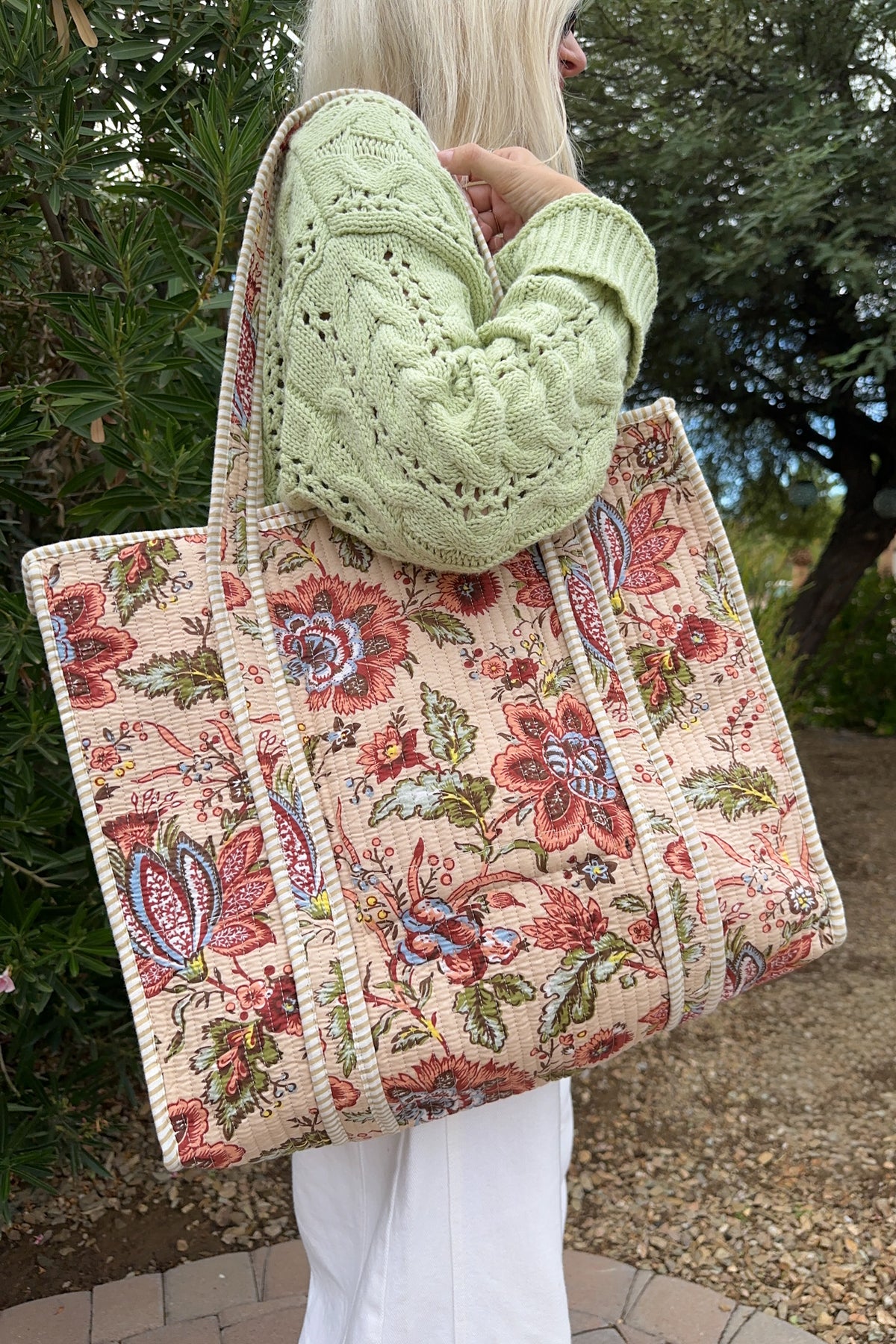 A person outdoors carrying a large woven tote bag with a floral and paisley pattern in pink, red, green, and blue on a cream background. The person is wearing a light green cable-knit sweater and white pants.