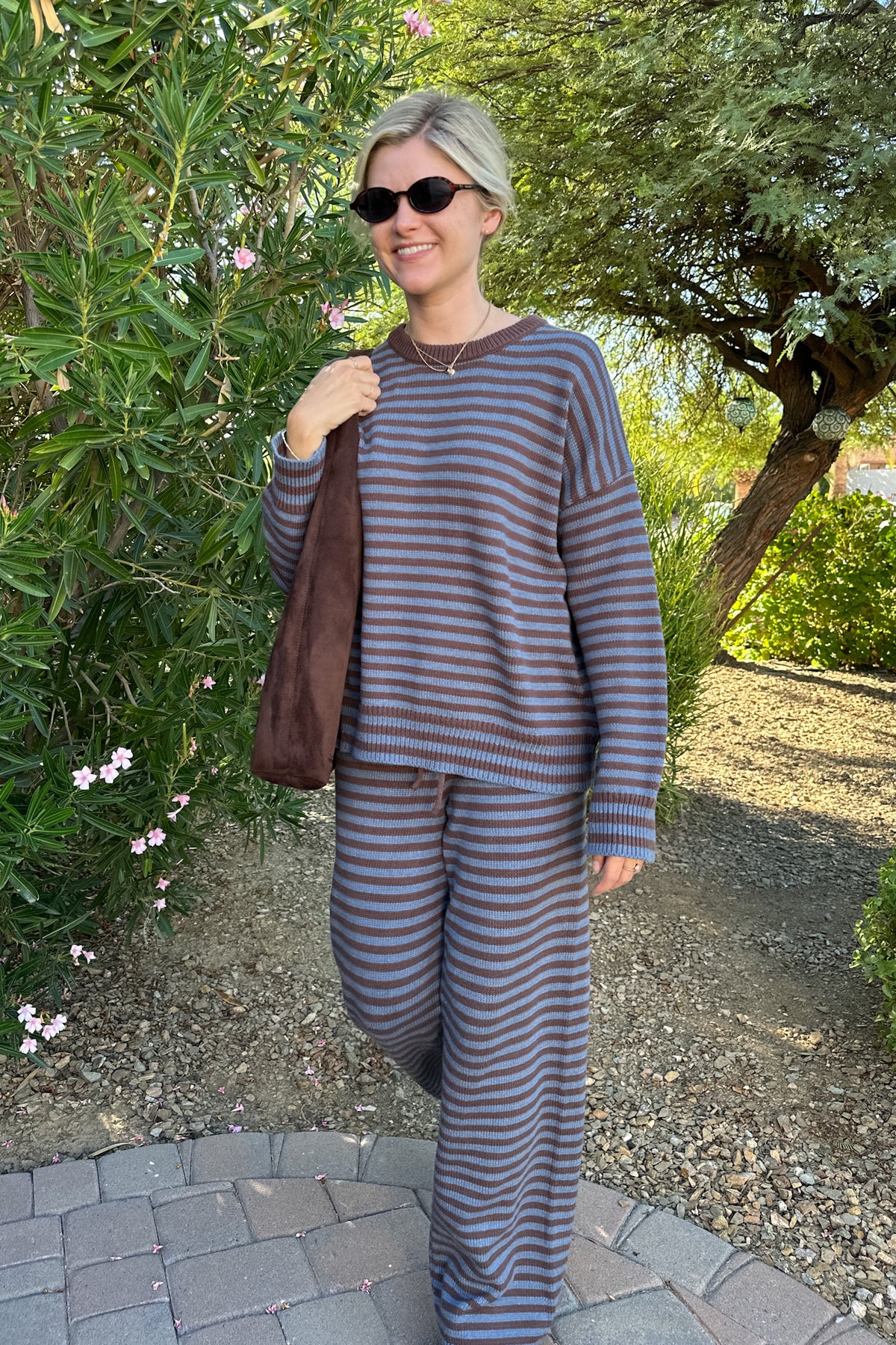 A woman wearing a matching two-piece lounge set with horizontal gray and brown stripes, standing outdoors on a stone path with green foliage in the background.