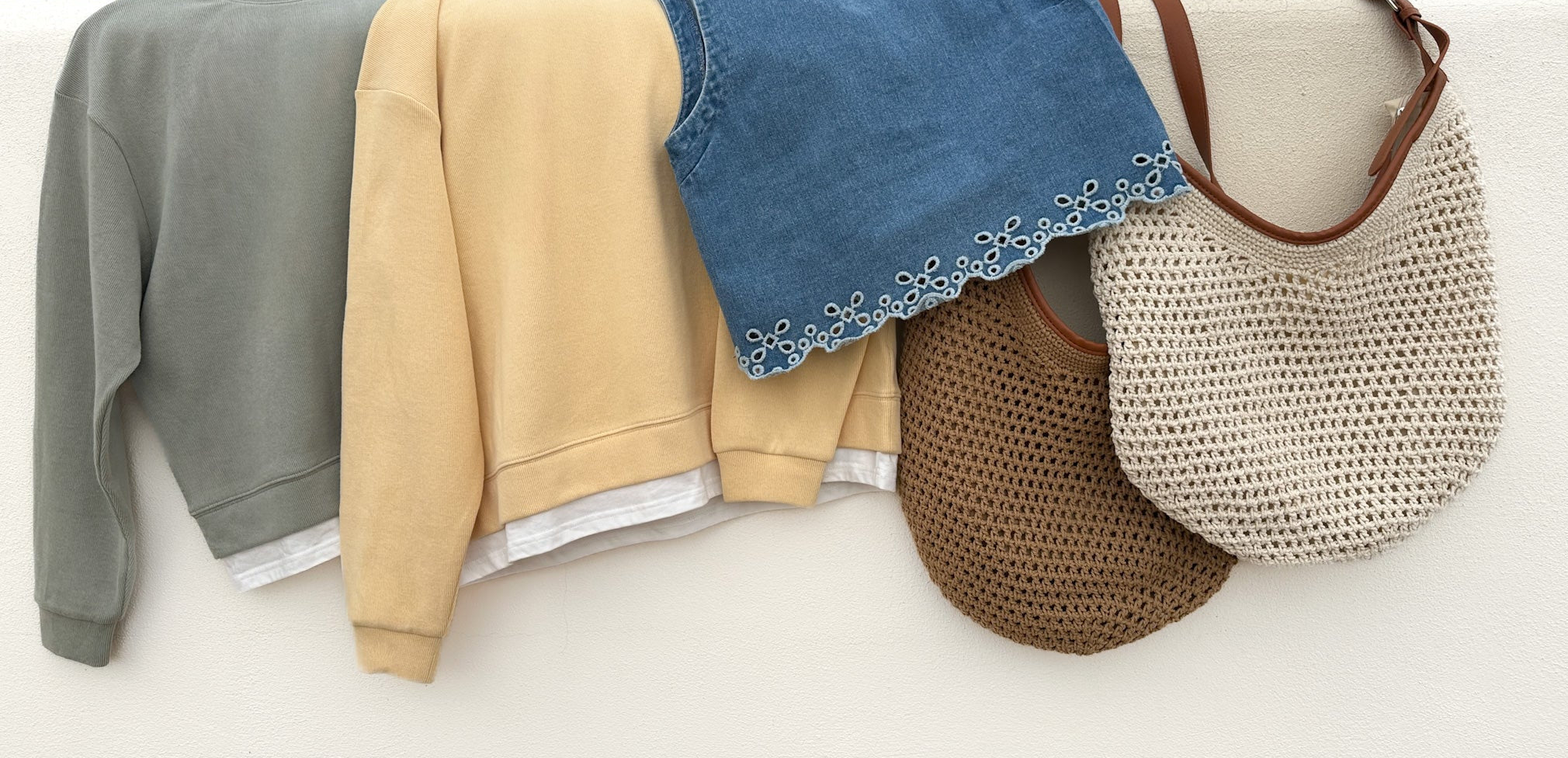 Stack of folded clothes including a gray sweatshirt, beige sweater, blue denim jacket, and two bags on a white surface.