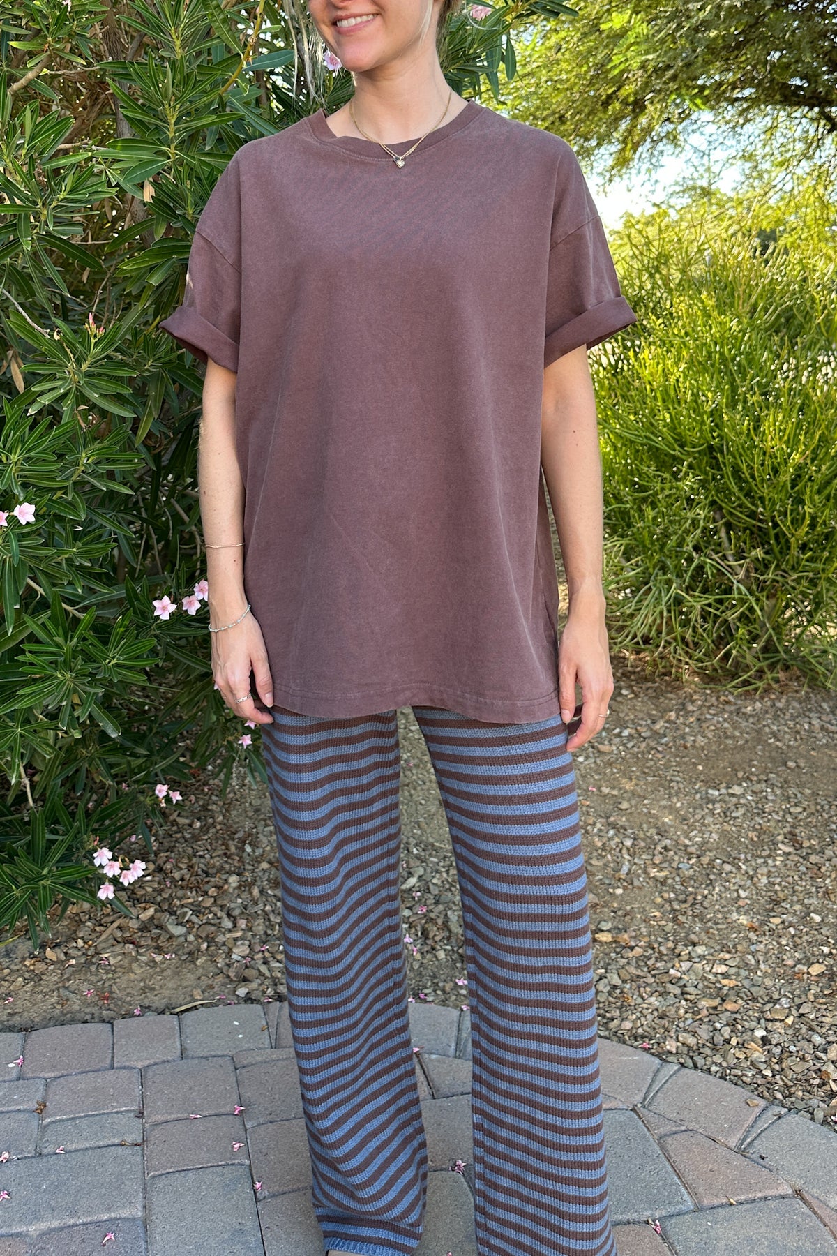 A woman wearing an oversized, short-sleeve crewneck t-shirt in a washed, muted mauve-brown color, paired with blue and gray striped wide-leg pants, standing outdoors on a paved path with green bushes in the background.