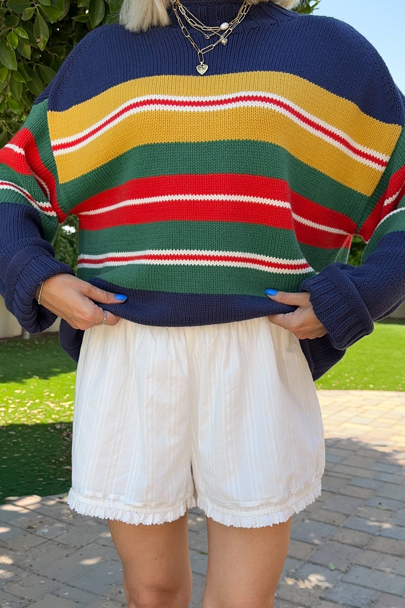 A person outdoors wearing a colorful striped knit sweater with navy, yellow, green, red, and white horizontal bands, paired with white shorts.