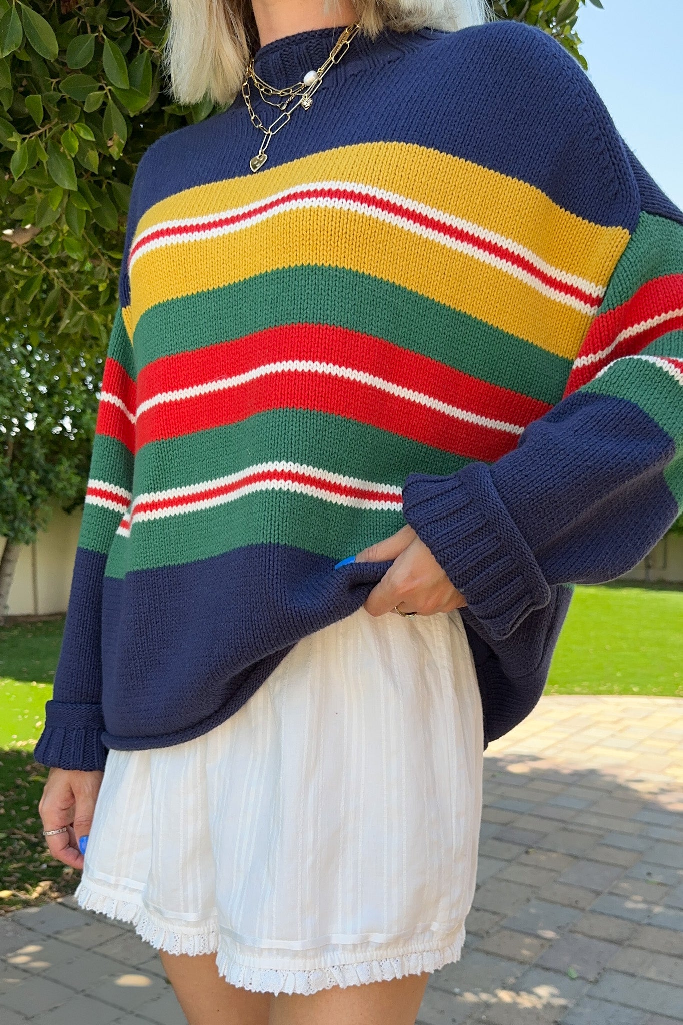 A person outdoors wearing a chunky knit sweater with bold horizontal stripes in navy, yellow, green, red, and white, paired with white shorts.