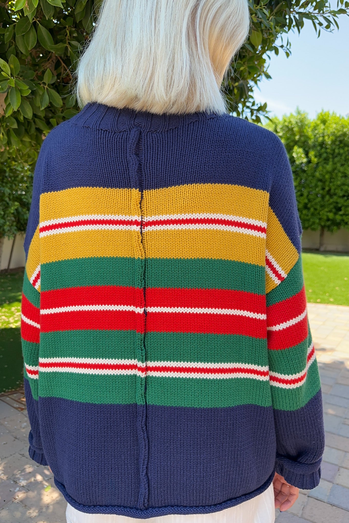 A person viewed from behind wearing a navy blue knit cardigan with wide horizontal stripes in yellow, green, red, and white, photographed outdoors on a paved walkway with greenery in the background.