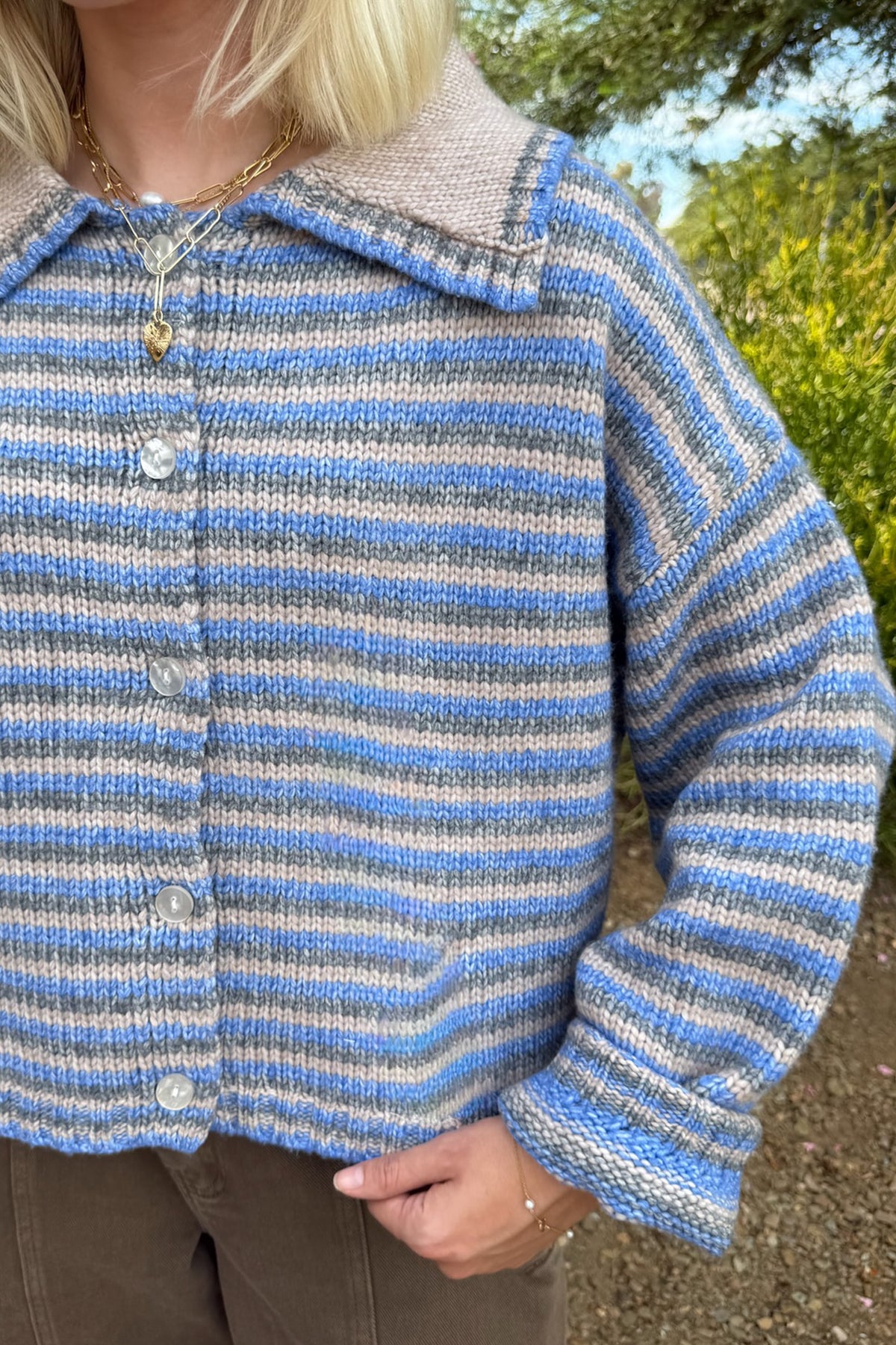 A close-up of a person wearing a hand-knit striped cardigan with blue, gray, and white horizontal stripes, featuring a beige knit collar and button closure.