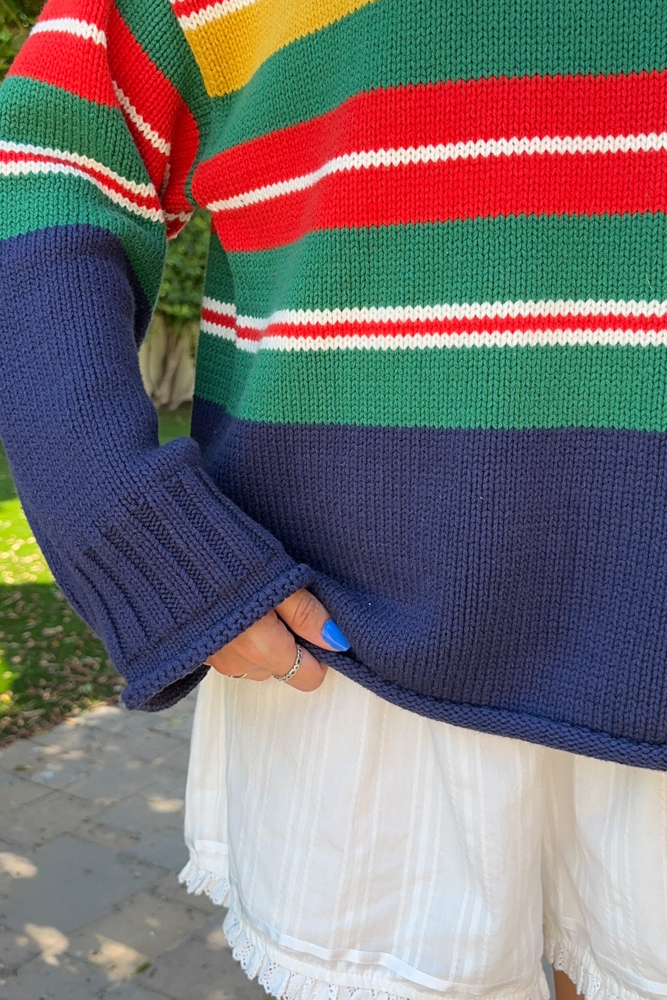 A close-up of a person wearing a colorful striped knit sweater with navy blue sleeves and a green, red, yellow, and white striped body, paired with white shorts outdoors.