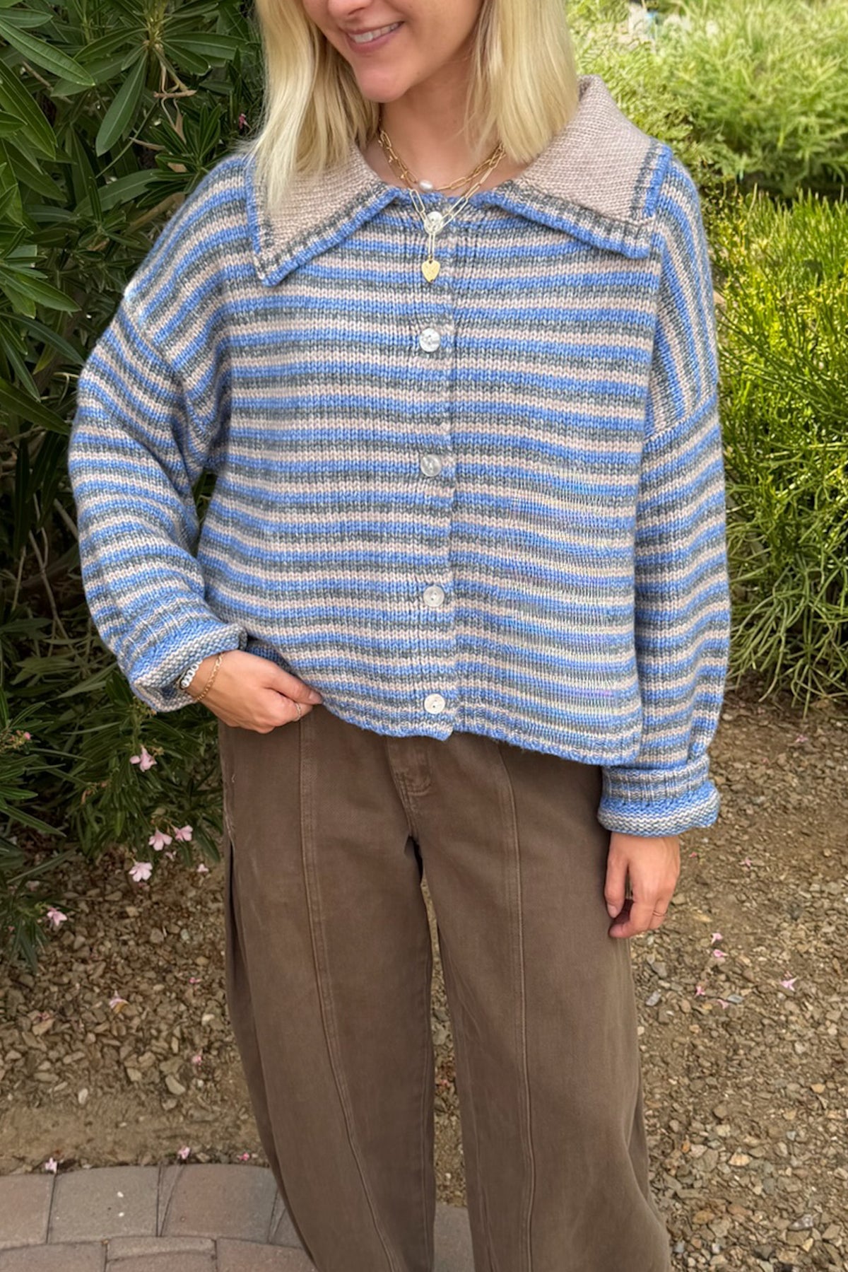 A woman wearing a blue and white striped knit cardigan with a beige collar, paired with brown trousers, standing outdoors in front of green garden foliage.
