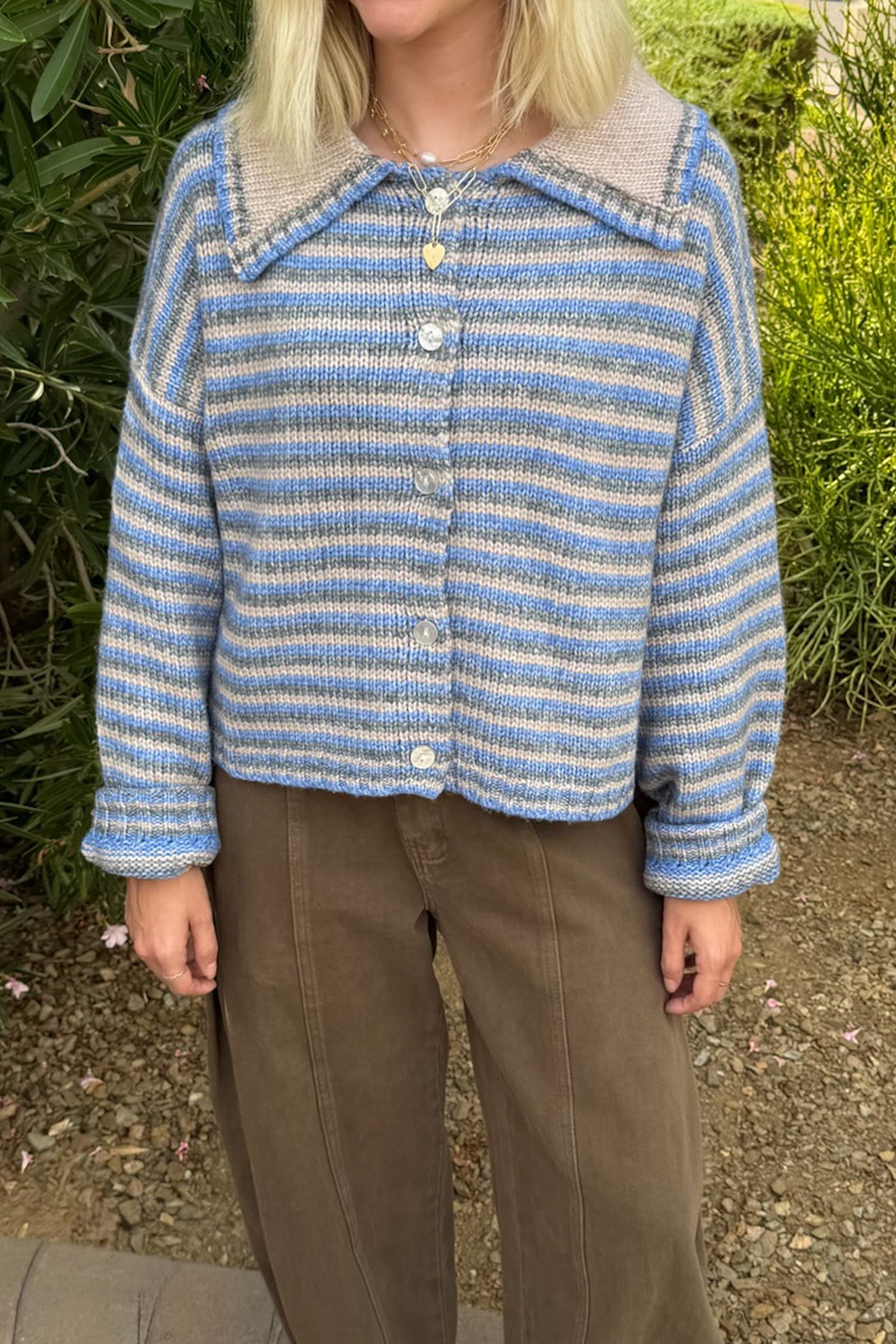 A person wearing a cropped, long-sleeve knit cardigan with horizontal blue, white, and beige stripes, featuring a soft beige collar and front buttons, paired with brown trousers, set against a background of green garden foliage.