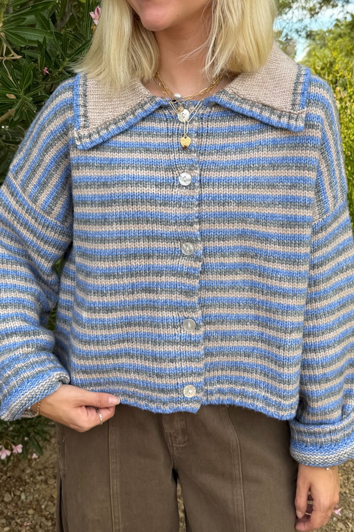 A person wearing a cropped knit cardigan with horizontal blue, beige, and gray stripes, featuring a beige knit collar and front buttons, paired with brown pants in an outdoor setting.