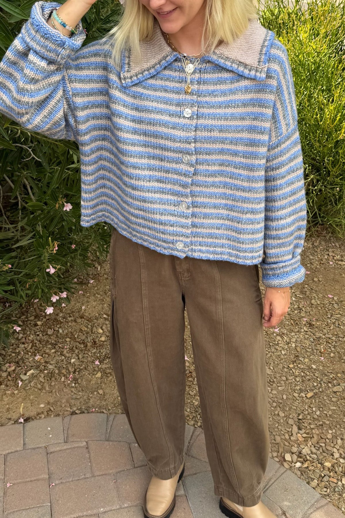A person wearing a cropped, long-sleeve knit cardigan with blue and beige horizontal stripes, a beige collar, and white buttons, paired with brown wide-leg pants and beige boots, standing outdoors on a paved path with green foliage in the background.