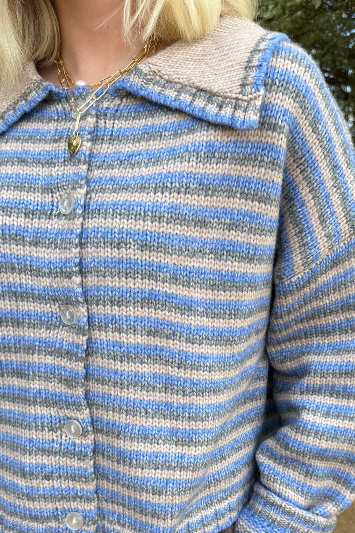 A close-up of a person wearing a hand-knit collared cardigan with horizontal stripes in light blue, gray, and off-white. The cardigan features a soft knit collar and a button-up front.