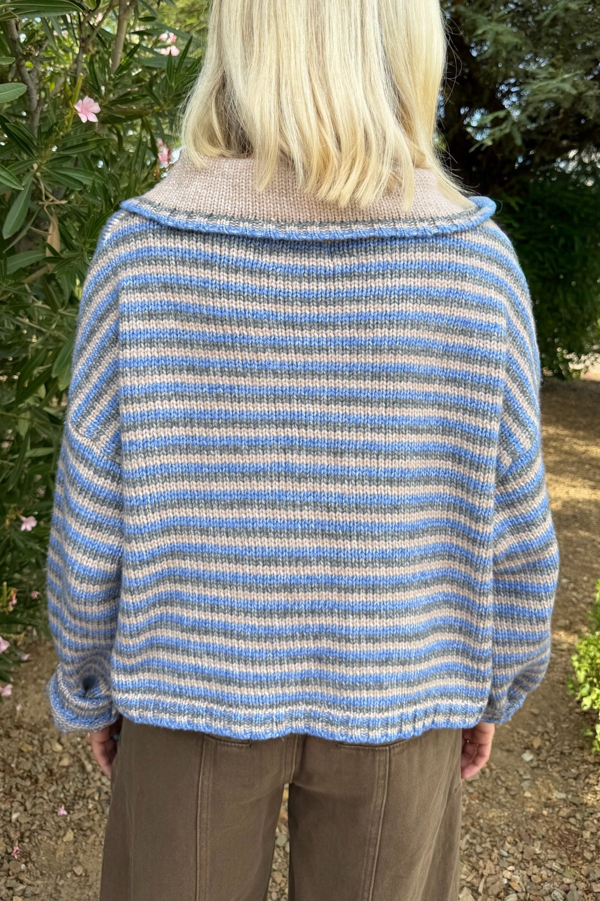 A person viewed from behind wearing a loose striped knit sweater outdoors. The sweater has horizontal stripes in light blue and off-white/cream, with a soft beige collar.