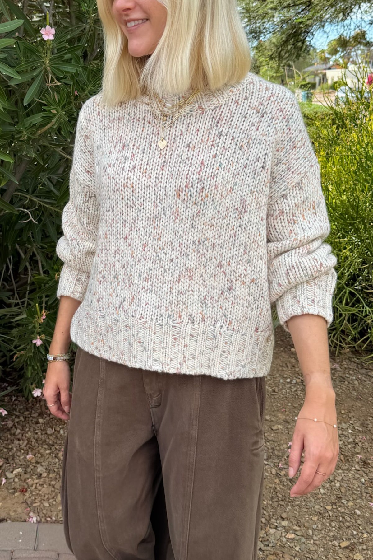 A woman wearing a chunky knit long-sleeve sweater in a cream and beige heathered pattern with subtle pink and brown flecks, paired with brown pants, standing outdoors in front of green garden foliage.