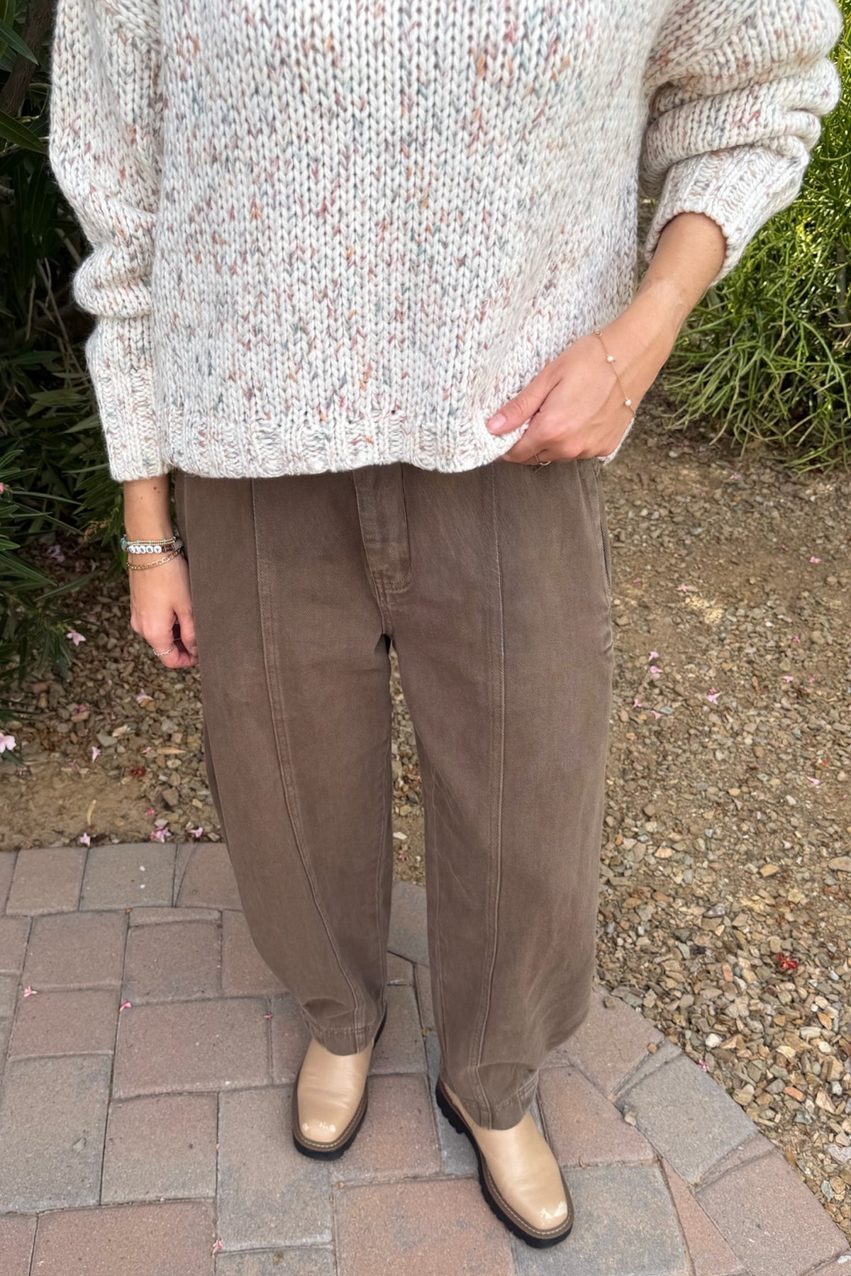 A person standing outdoors on a brick path wearing a chunky knit sweater, loose brown pants, and beige shoes.