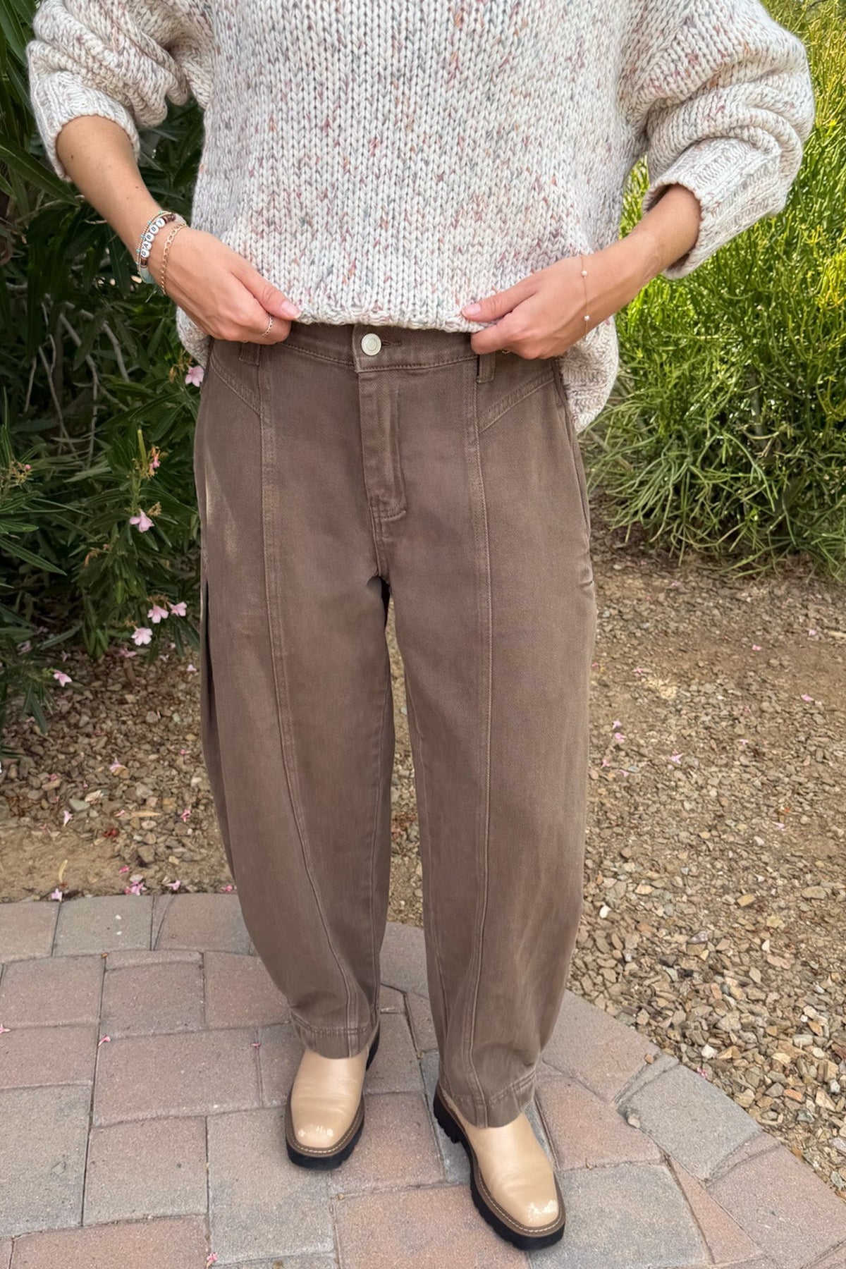 A person standing outdoors on a brick patio wearing loose brown wide-leg pants, a cream knit sweater, and beige shoes, with greenery and gravel in the background.