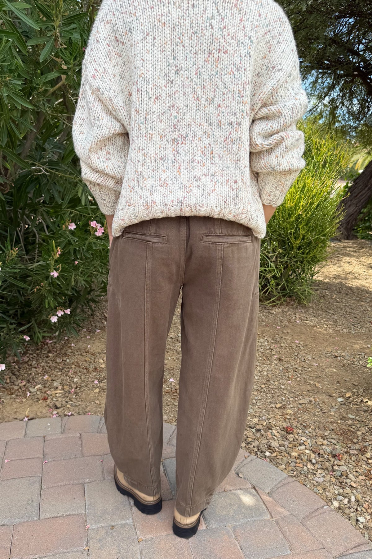 A person standing outdoors on a brick path, viewed from the back, wearing a cream knit sweater with tiny multicolor flecks and brown wide-leg pants.